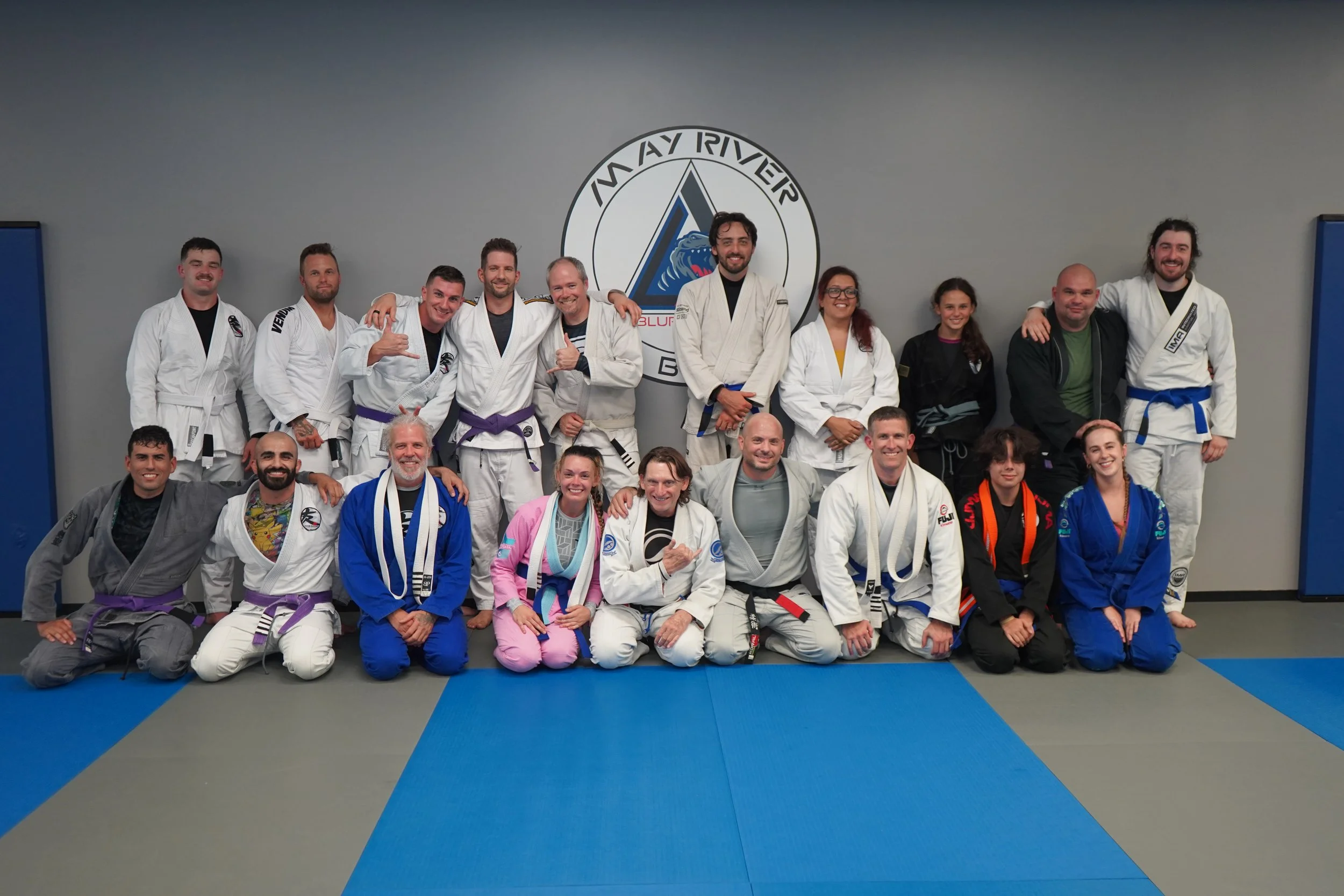 Group of people in Brazilian Jiu-Jitsu uniforms posing on mats in a martial arts gym.
