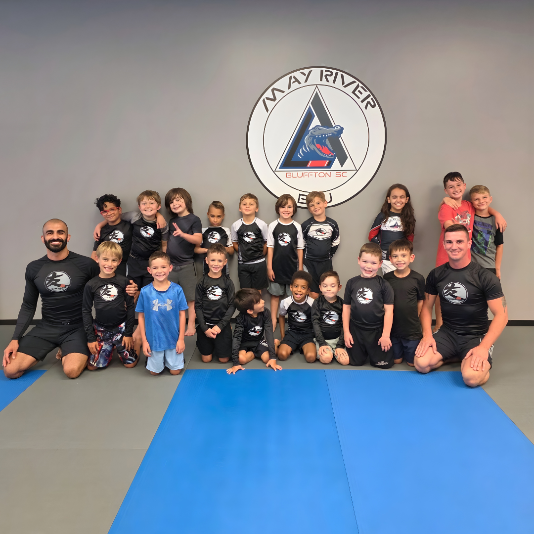 Group of children and two adult instructors in a martial arts dojo, with a May River Martial Arts logo on the wall behind them.