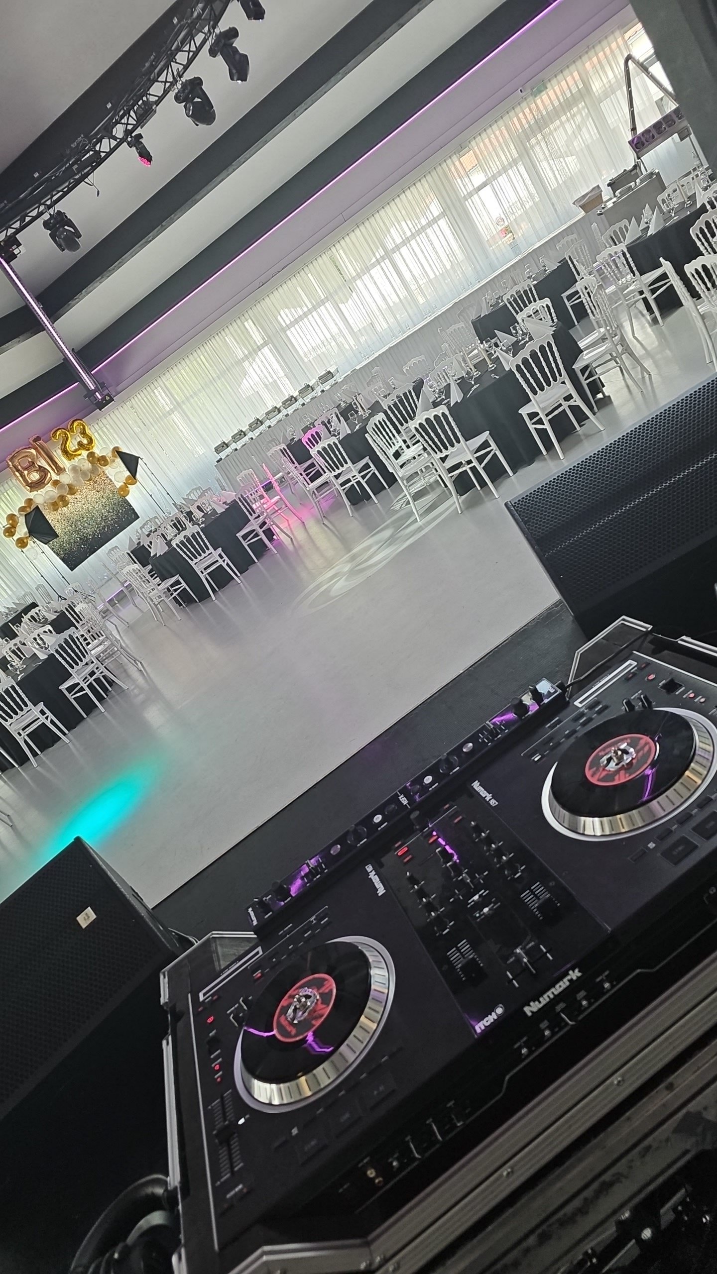 Empty event hall decorated with black tablecloths and white chairs, with a dance floor and a DJ booth in the foreground, and balloon decorations and a '2023' sign on one wall.