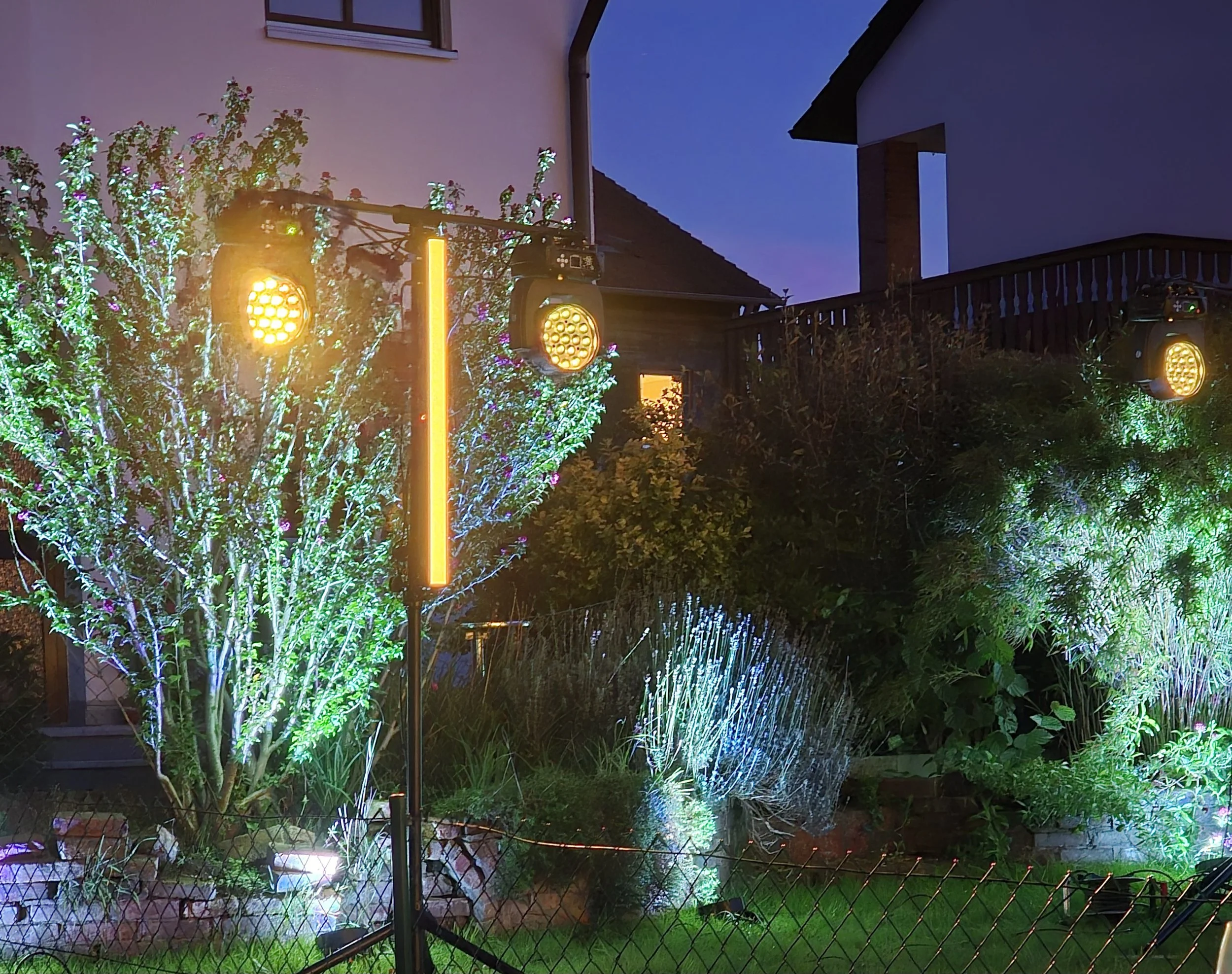 Outdoor garden scene at night with colorful LED lights illuminating trees and bushes, and stage lighting equipment set up in the garden.