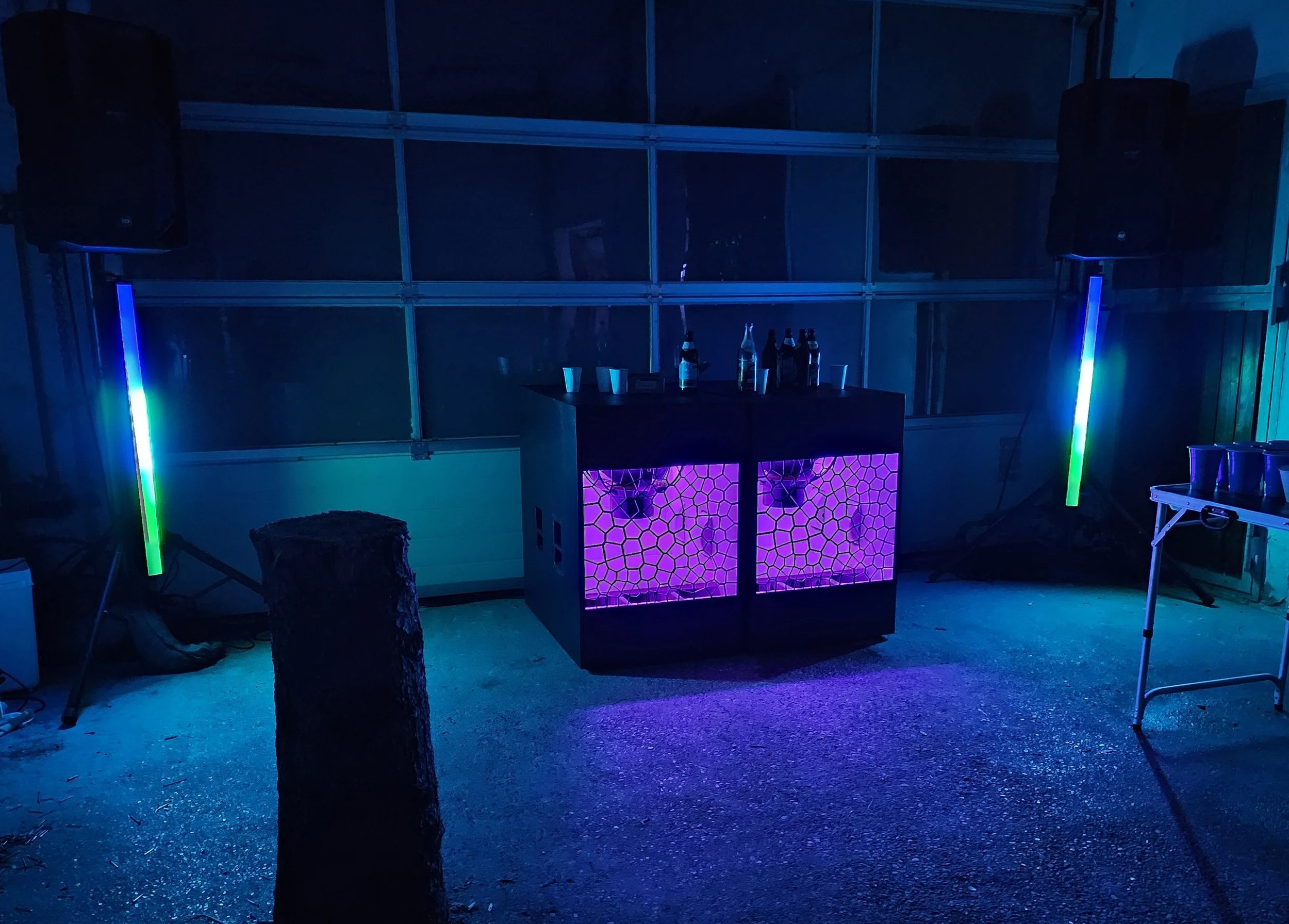 A DJ booth with pink glowing mesh panels and bottles on top, illuminated by colorful LED lights in a dark setting.