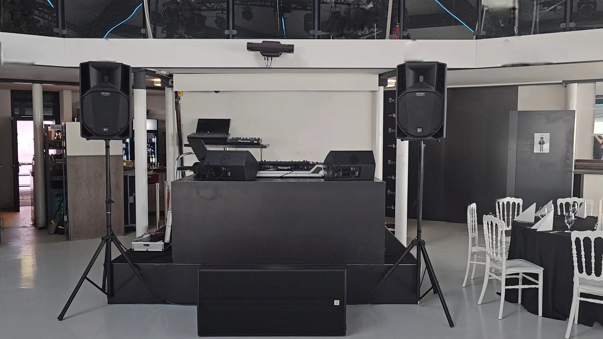Wedding reception area with DJ setup including speakers, DJ equipment on a black table, and seating with tables and white chairs.