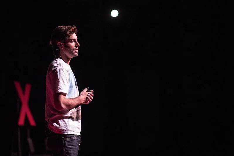 Alex Regen speaking at a TEDx talk, presenting on stage to a live audience