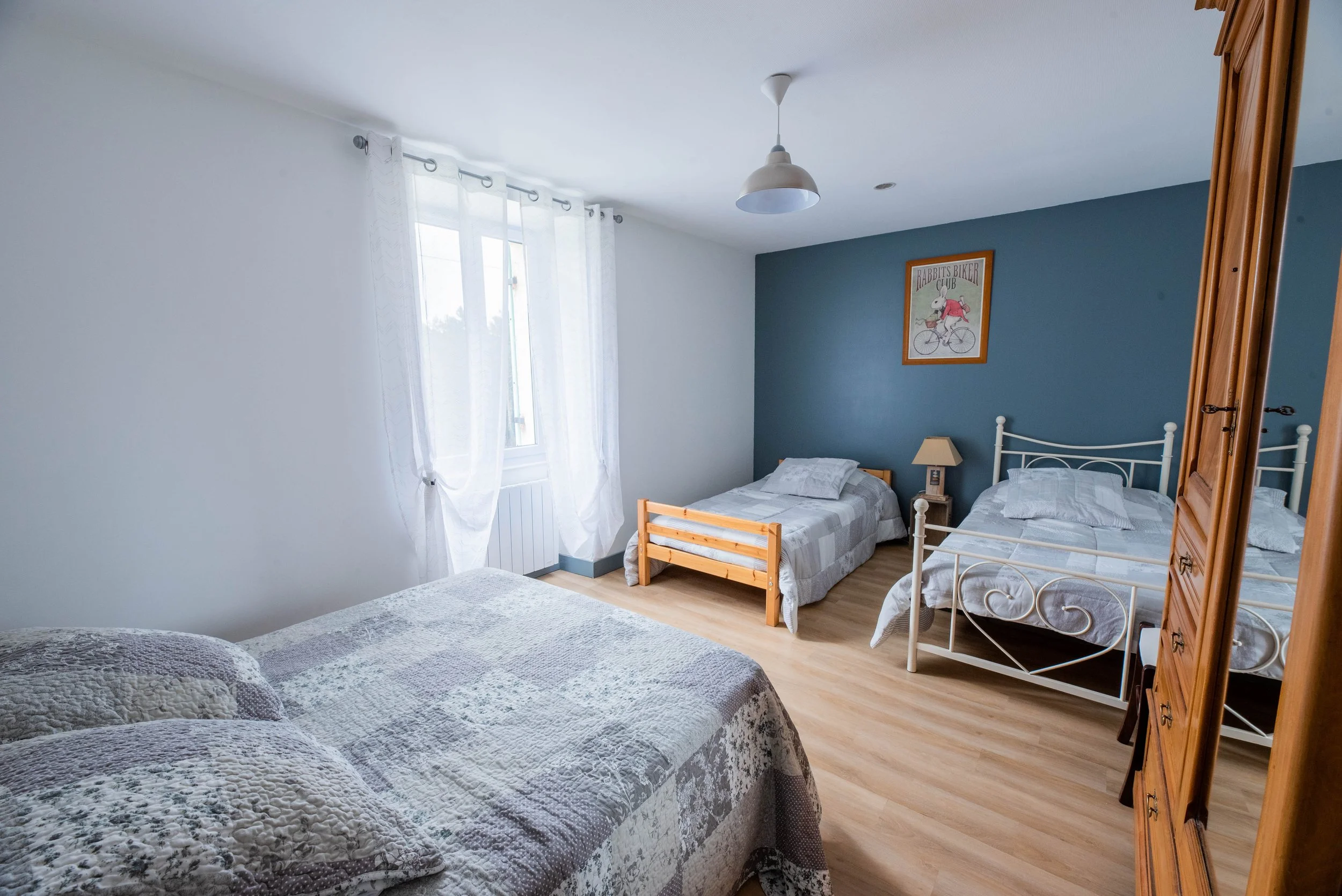 Gite Teulet Dordogne. Chambre avec trois lits, deux petits et un double, fenêtre avec rideaux blancs, mur bleu derrière les lits, tableau avec un lapin à vélo, armoire en bois, lampe de chevet, sol en bois clair.
