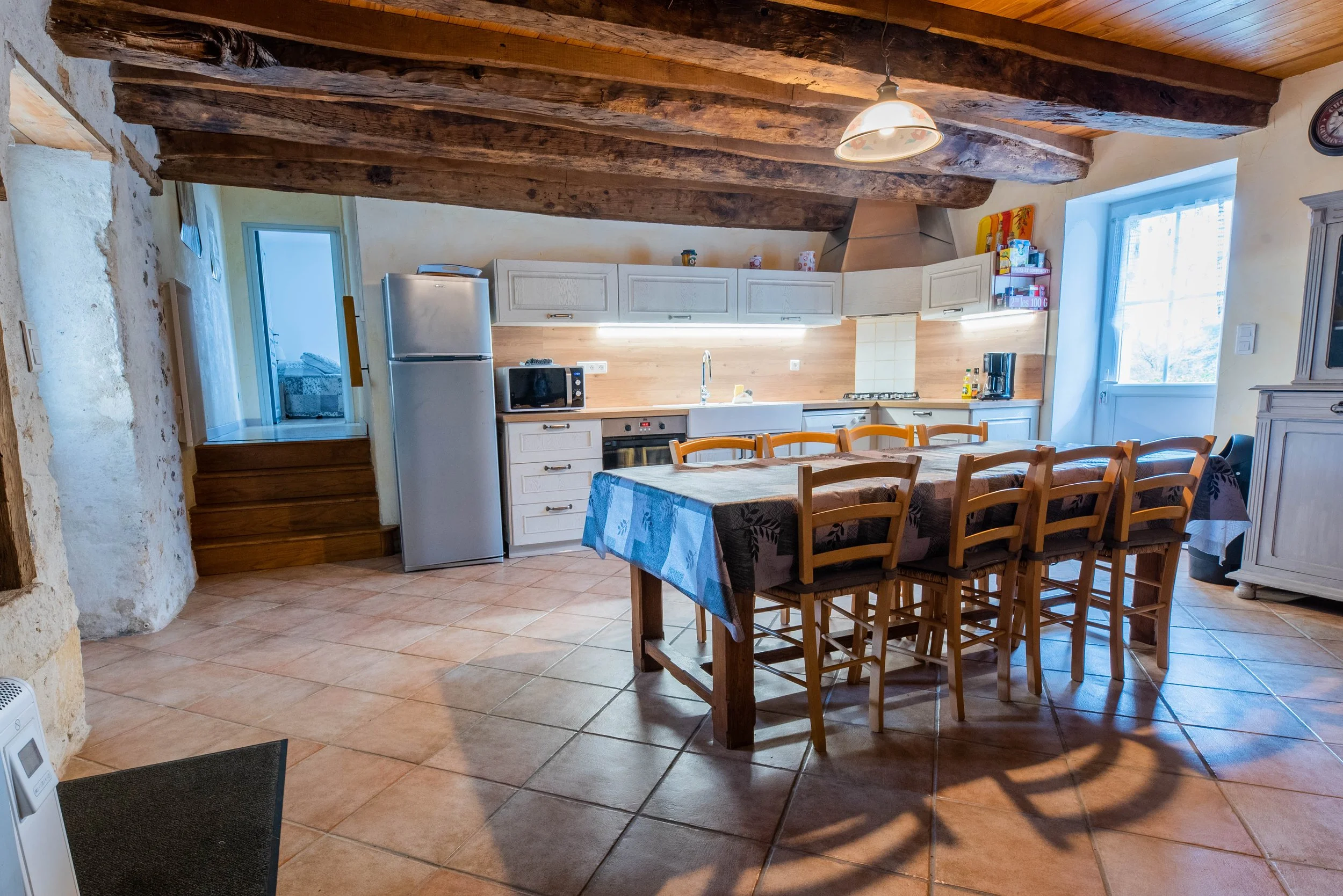 Gite Teulet Dordogne. Cuisine rustique avec poutres en bois, table à manger, chaises en bois, réfrigérateur, micro-ondes, évier, fenêtre lumineuse, décor champêtre.