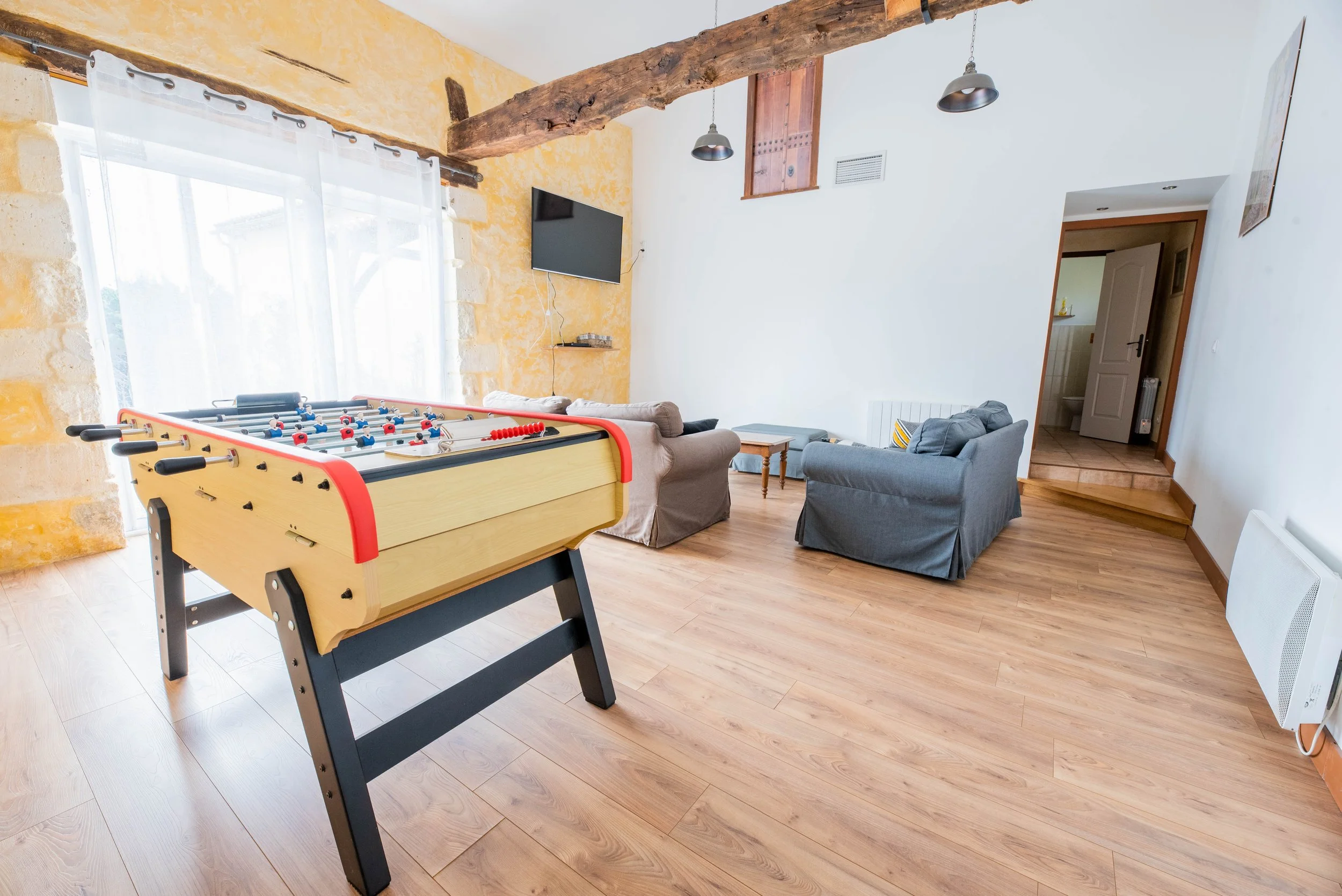 Gite Teulet Dordogne. Salon avec un baby-foot en bois, deux canapés gris, une télévision, une fenêtre avec rideaux blancs, un mur en pierre jaune, un parquet en bois, un petit escalier en bois, une porte entrouverte, et un radiateur blanc.