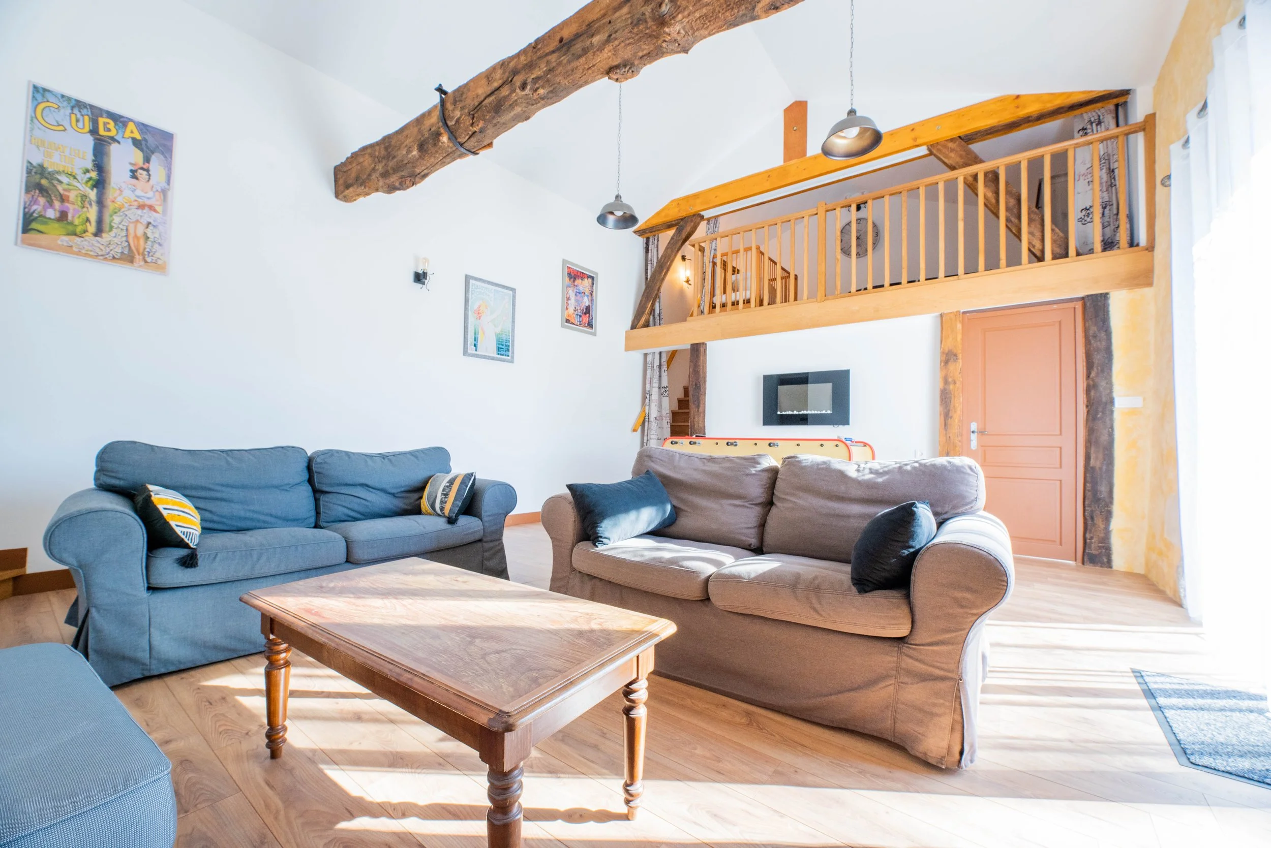 Gite Teulet Dordogne. Salon lumineux avec deux canapés, table en bois, murs décorés avec affiches, mezzanine en bois avec rampe, poutres en bois apparentes, porte en bois, et sol en parquet.