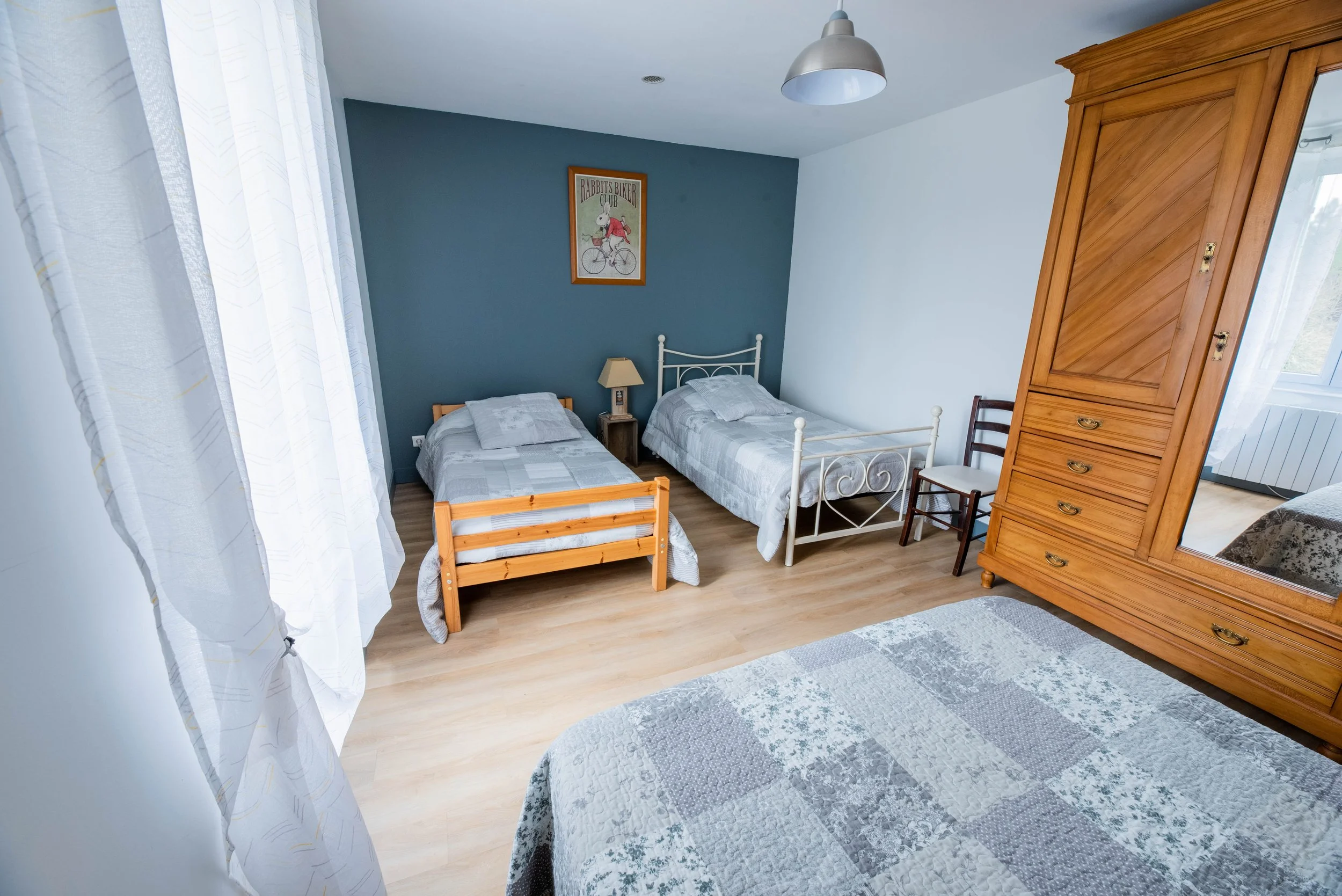 Gite Teulet Dordogne. Chambre avec deux lits simples, une armoire en bois, et un petit lit en bois, murs peints en blanc et bleu, fenêtre avec rideaux blancs, tableau d'un cycliste dans un cadre en bois.