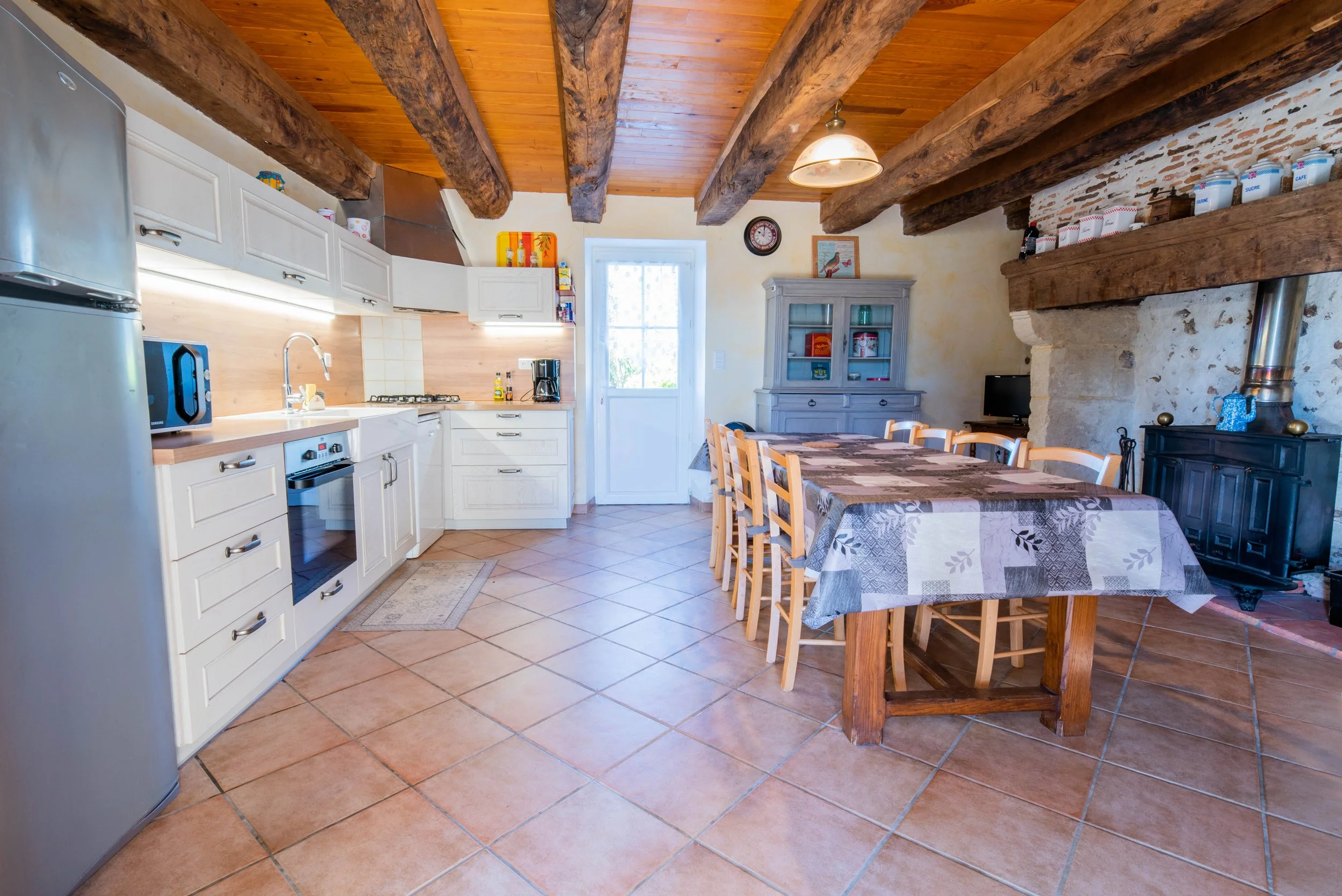 Gite Teulet DordogneCuisine de style rustique avec poutres en bois apparentes, comptoirs en bois, armoires blanches, table à manger en bois avec une nappe à motifs, cheminée en pierre avec un poêle, un meuble de rangement gris et un petit téléviseur.
