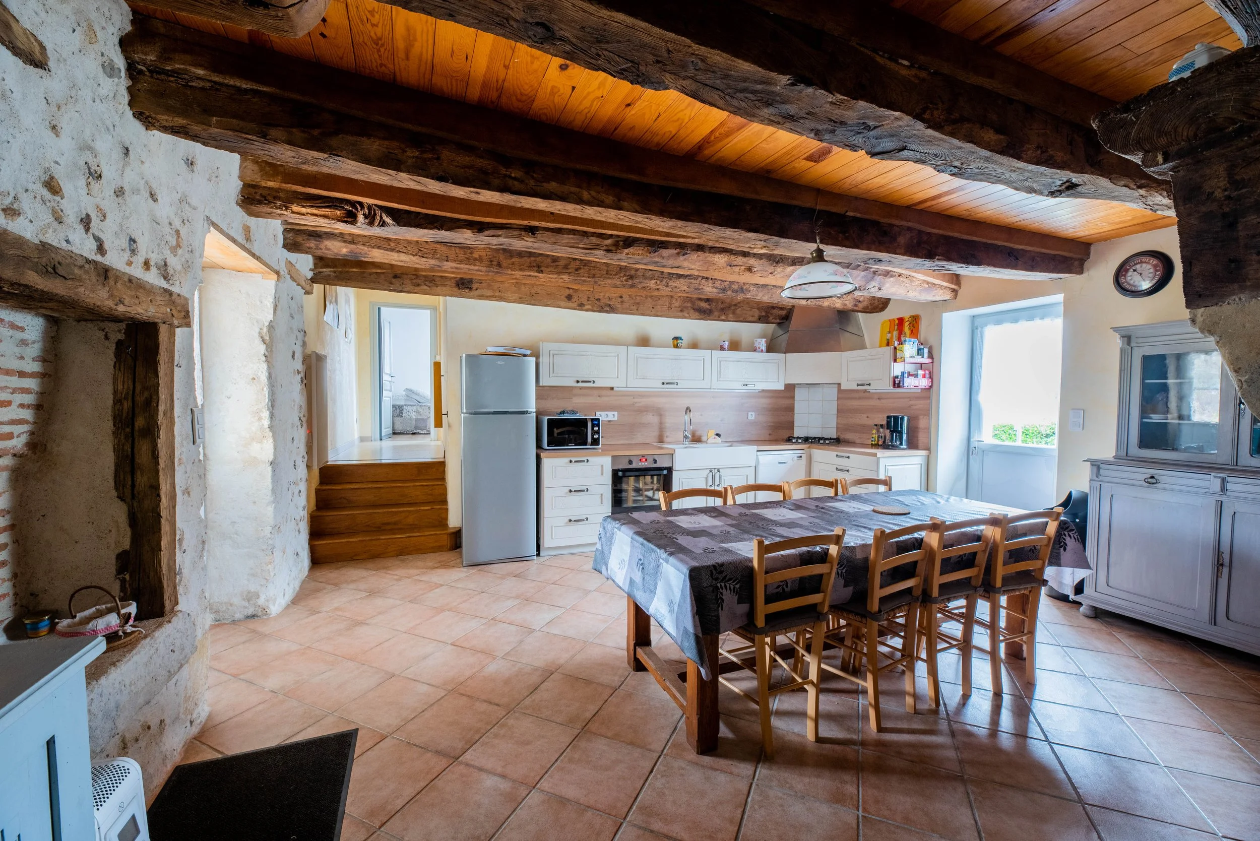 Gite Teulet Dordogne. Cuisine rustique avec poutres en bois apparentes, table en bois avec nappe, appareils électroménagers inox, armoires blanches, céramique beige au sol et lumière naturelle