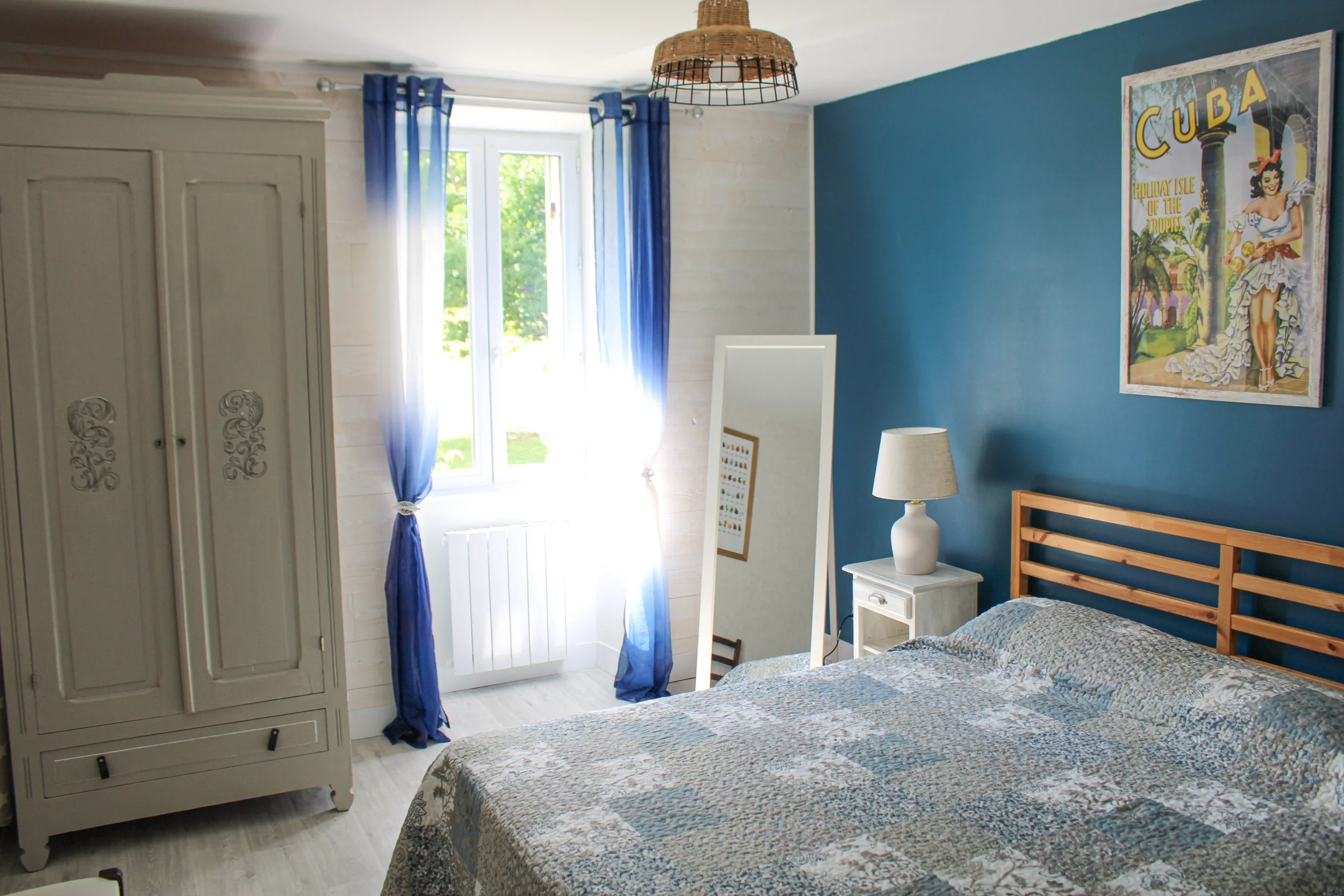 Gite Teulet Dordogne. Chambre à coucher lumineuse avec un lit, une armoire, une table de nuit, une lampe et un miroir, murs bleu et blanc, fenêtre avec rideaux bleus laissant entrer la lumière, affiche de Cuba au mur.