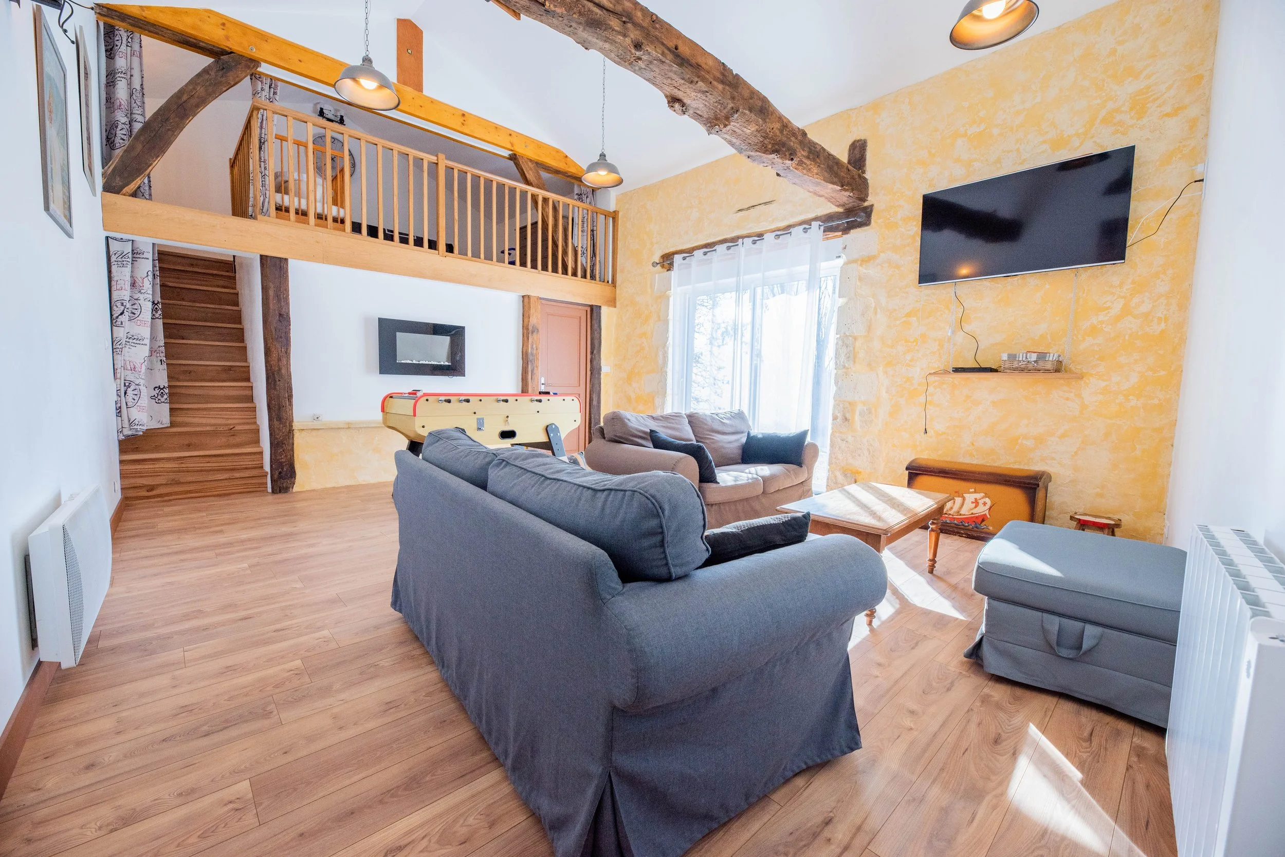 Gite Teulet Dordogne. Salon avec canapé gris, fauteuil beige, table basse en bois, TV à écran plat sur un mur jaune, grande fenêtre avec rideaux blancs, escalier en bois, mezzanine avec rampe en bois, lampes suspendues, sol en parquet