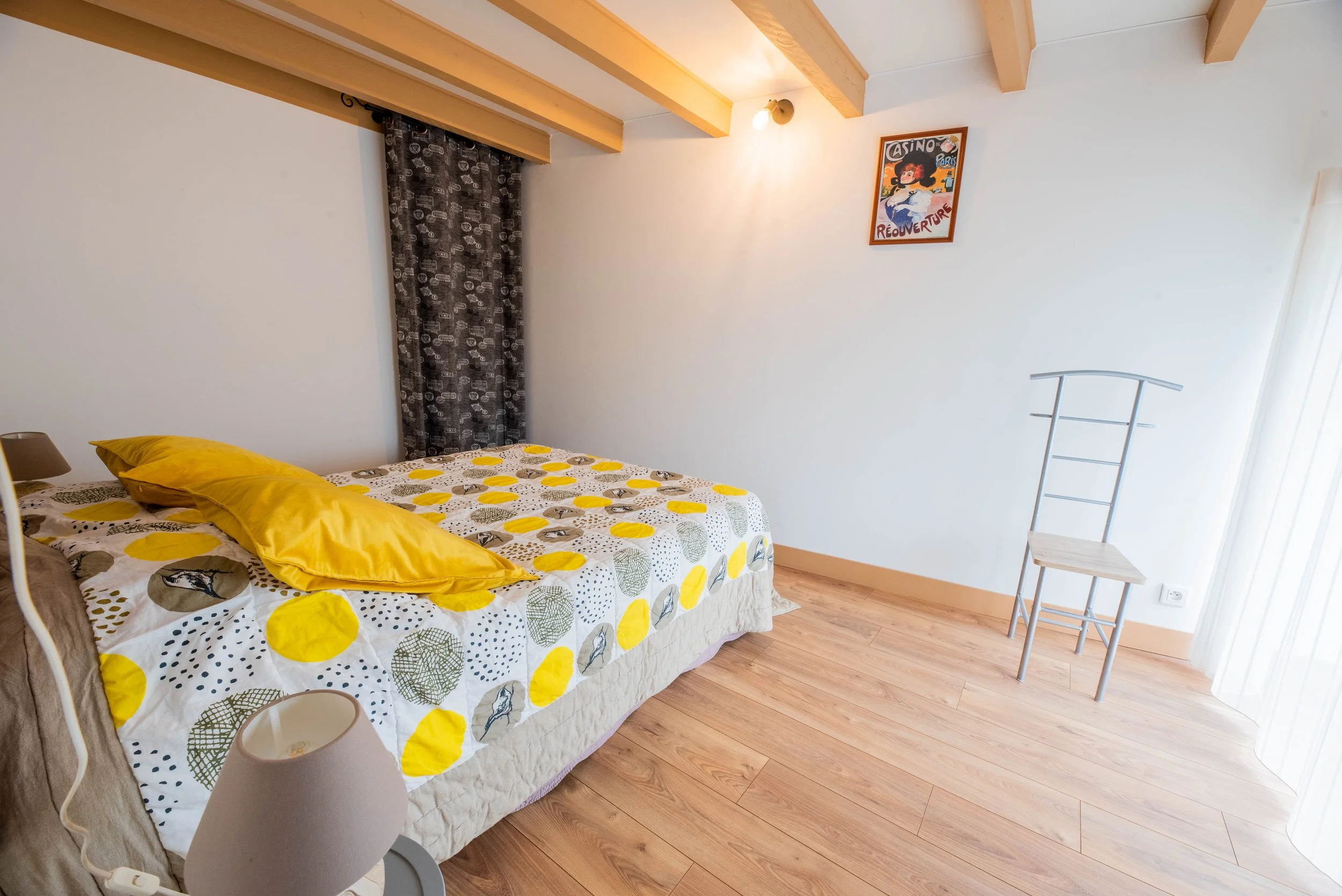 Gite Teulet Dordogne. Chambre avec lit, drap à motifs, oreillers jaunes, lampe de chevet, tableau sur le mur, chaise en bois, porte-fenêtre avec rideaux transparents, sol en bois, plafond avec poutres en bois.