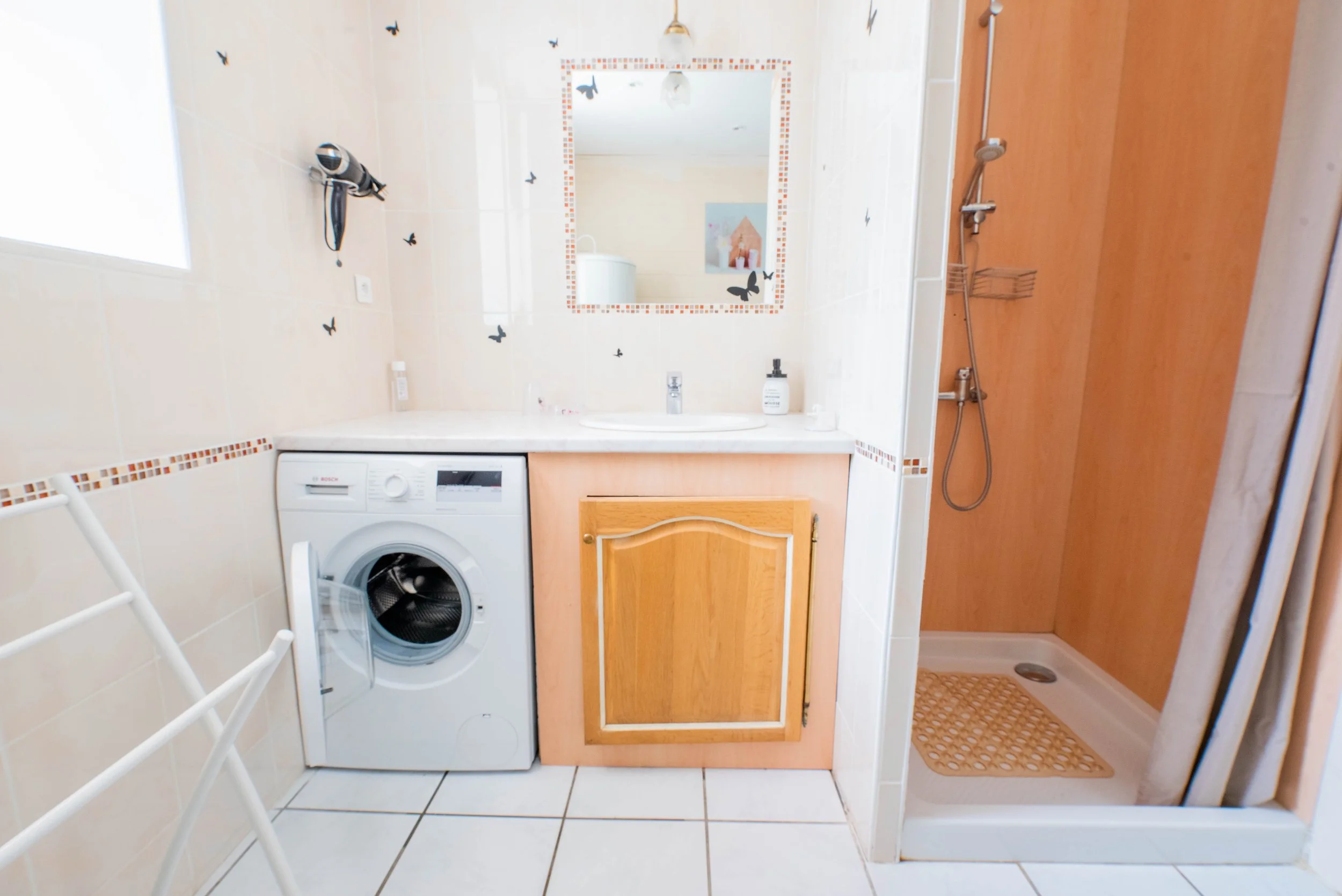 Gite Teulet Dordogne. Salle de bain avec lavabo, miroir décoré de papillons, machine à laver, armoire en bois, douche avec rideau, reflet d'une pièce avec peinture.