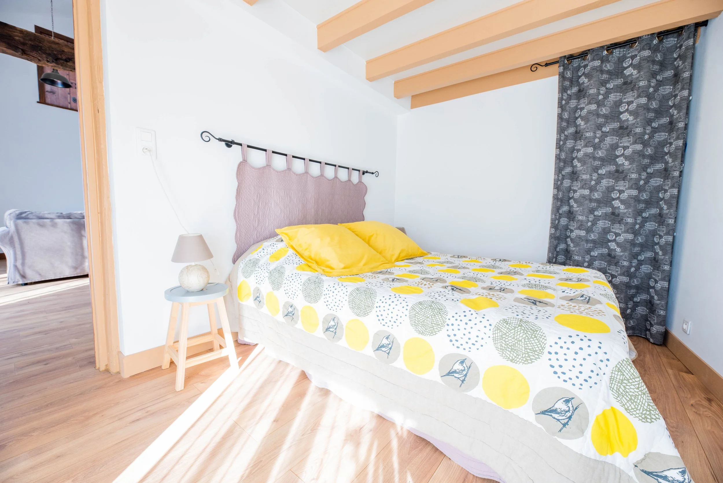Gite Teulet Dordogne. Chambre avec lit double, tête de lit mauve, drap à motifs verts, jaunes et gris, deux oreillers jaunes, petite table avec lampe, rideau gris à motifs, sol en bois clair, plafond avec poutres en bois.