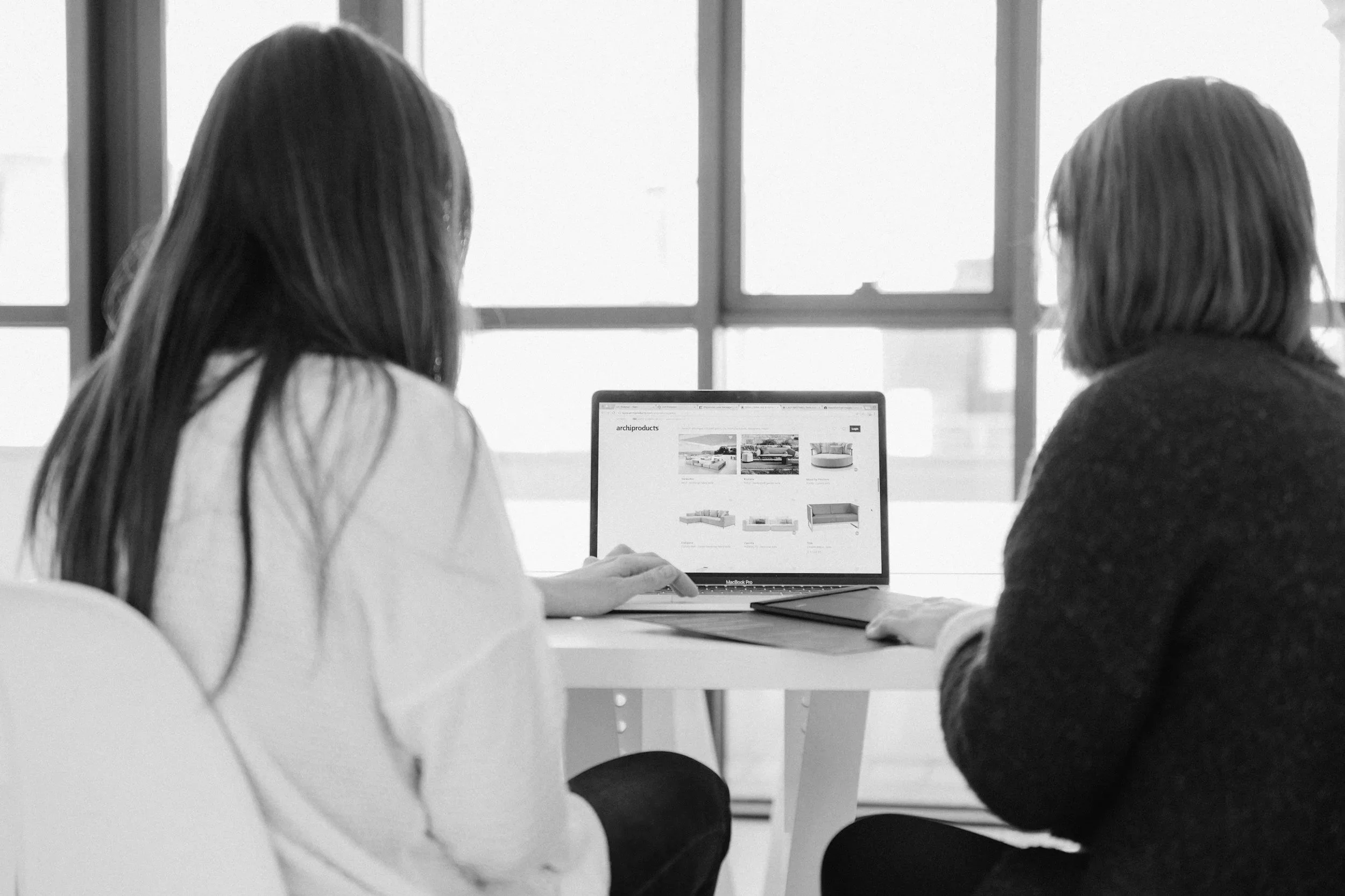 Two women side by side, both looking a one laptop discussing work related things.
