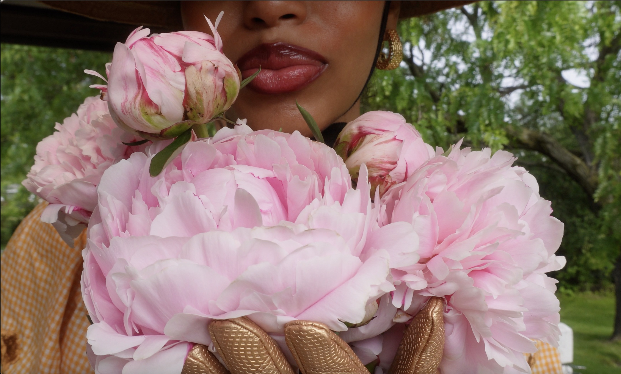 A person with dark lipstick and gold earrings holding a large pink peony flower close to their lips outdoors, with green trees in the background.