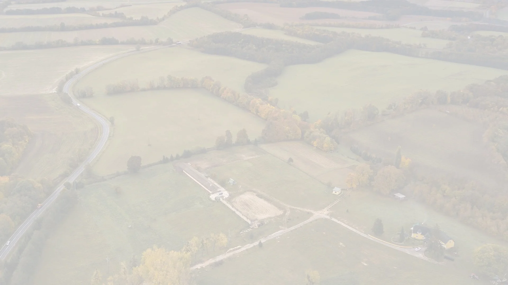 Aerial view of a land with fields, trees, a winding road, and a few houses.
