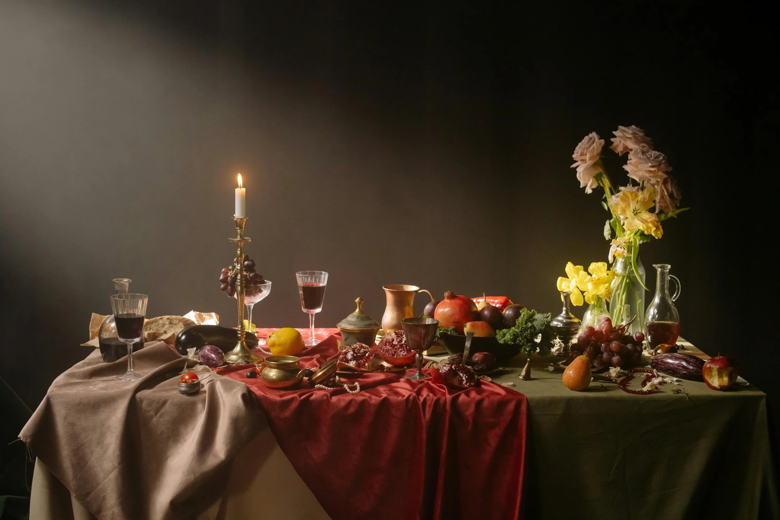 Una mesa decorada con frutas, vino, velas y flores en un fondo oscuro.