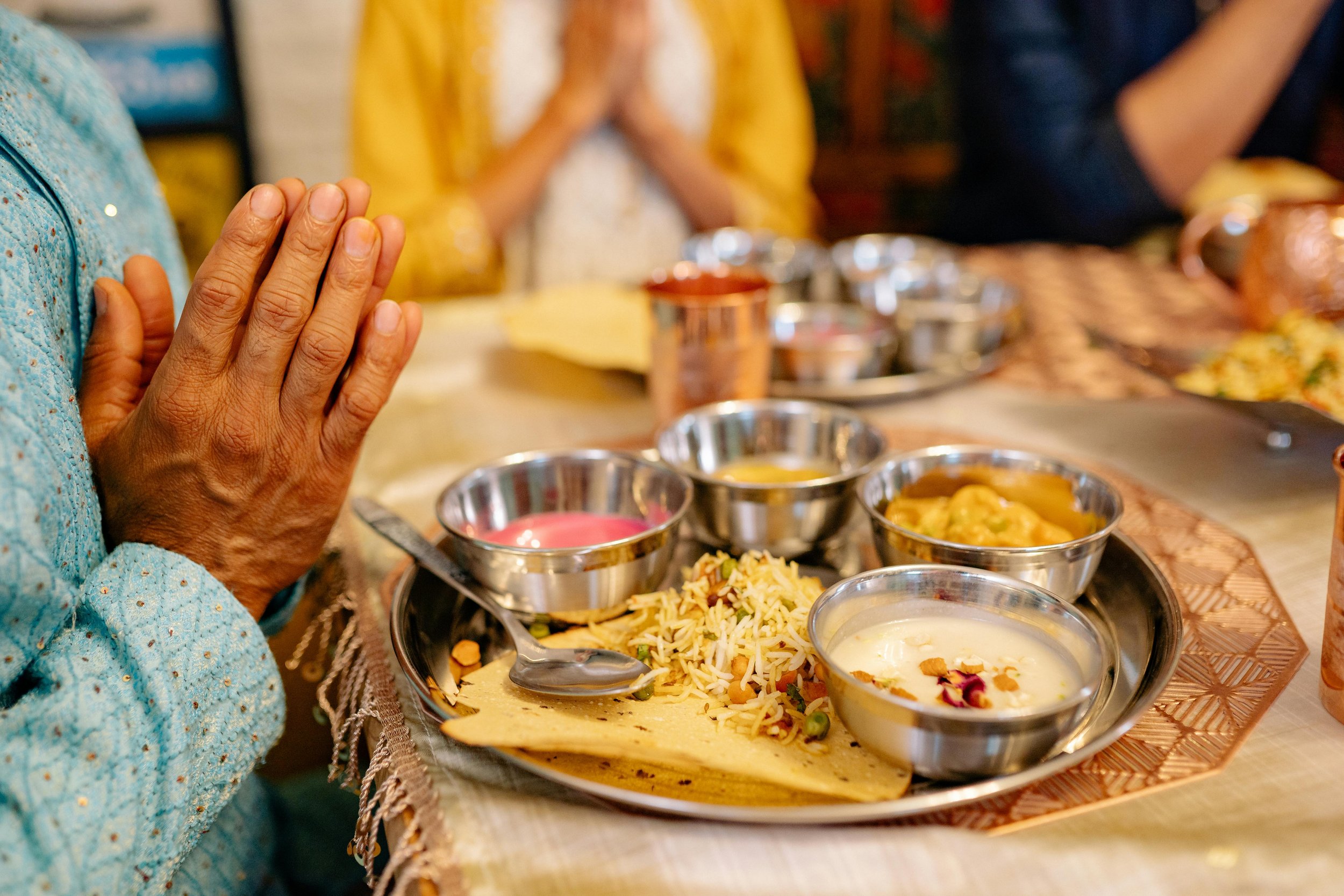 Manos en posición de oración en una comida tradicional india, con platillos de comida y utensilios en la mesa.