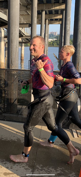 Two runners, a man and a woman, smiling and running barefoot along a riverside path near a bridge, holding water bottles.