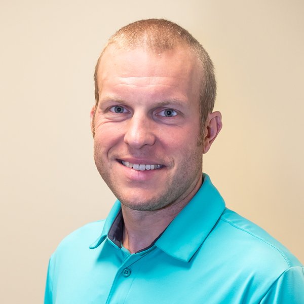A smiling man with short light brown hair and blue eyes, wearing a turquoise collared shirt, standing against a plain beige background.