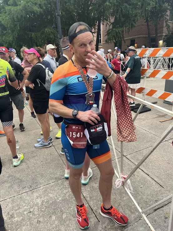 A man in colorful athletic gear after finishing a race, wearing a medal around his neck, holding a water bottle, and adjusting his cap, surrounded by other runners and race infrastructure.