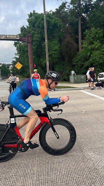 A cyclist wearing a multicolored jersey and helmet rides a black and red bike on a city street with pedestrians and trees in the background.