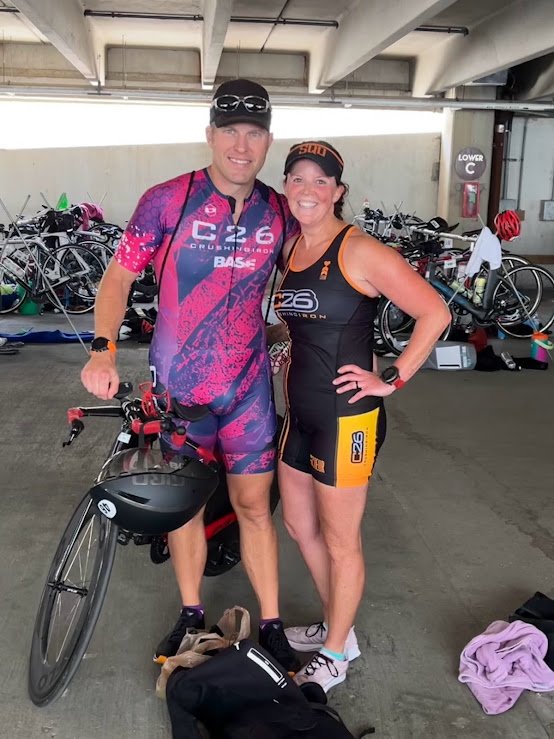 A man and woman in athletic gear smiling at a triathlon or cycling event, standing inside a parking garage with bicycles in the background.