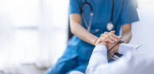 A healthcare professional in blue scrubs holding a patient's hand in a medical setting.