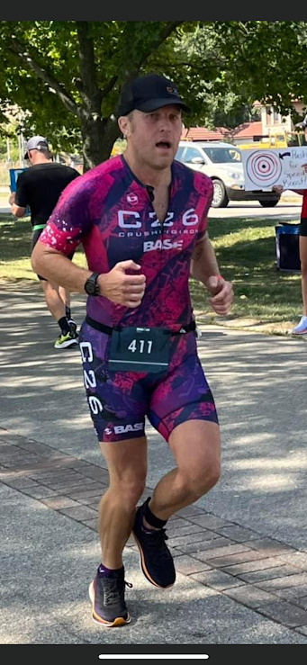 A male runner wearing a pink and purple athletic outfit, black cap, and running shoes, participating in a race outdoors with green trees, other runners, and event tents in the background.