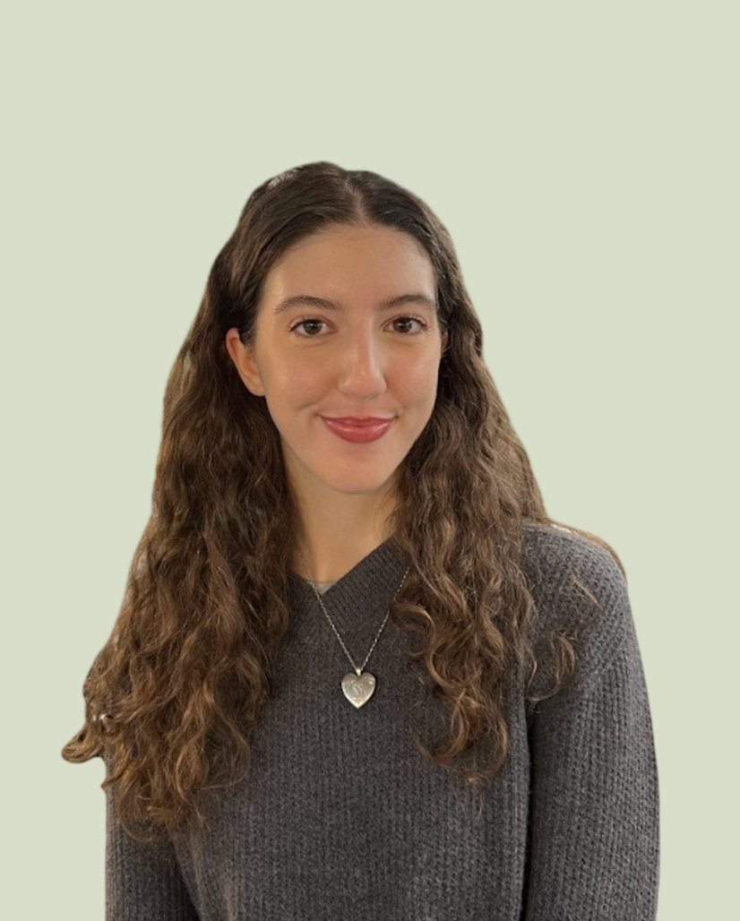 A woman with long, wavy brown hair and a light complexion, wearing a dark gray sweater and a silver heart-shaped necklace, smiling at the camera against a pale green background.