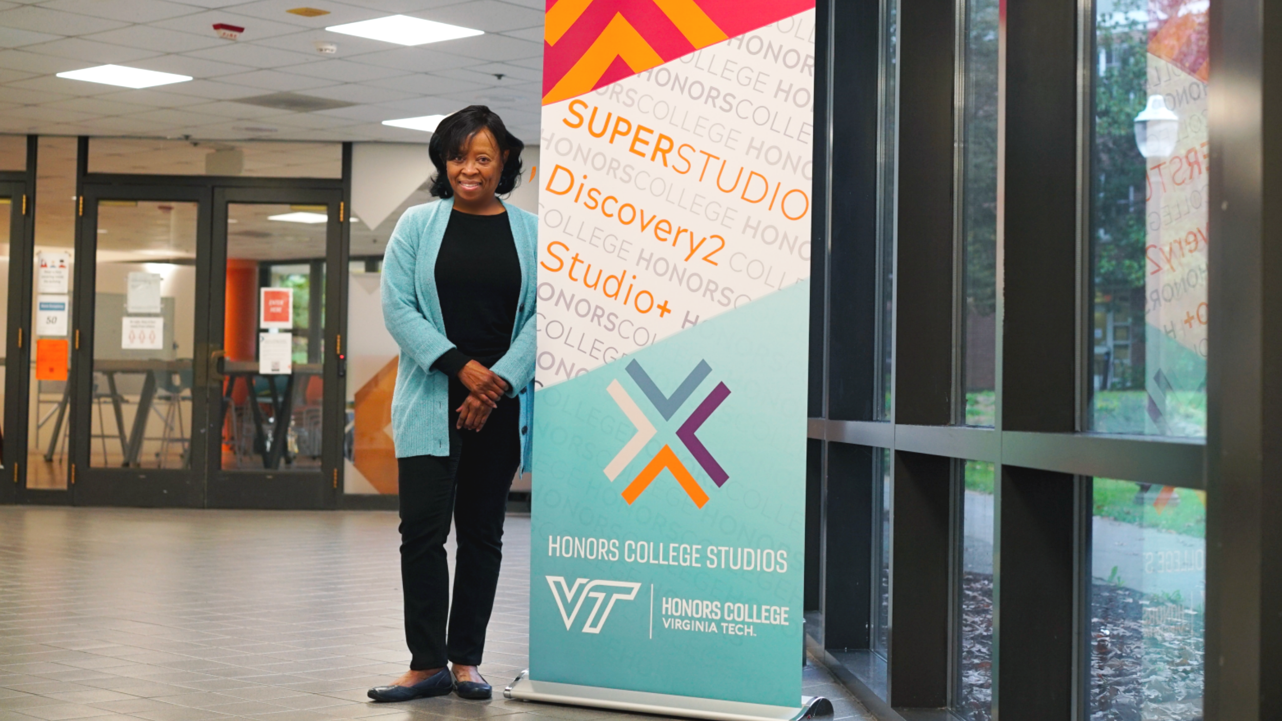 A woman standing indoors next to a Virginia Tech Honors College Studies banner, smiling and dressed in a black top, blue cardigan, black pants, and black shoes.
