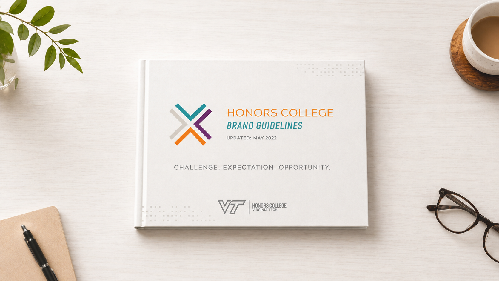 A white book titled 'Honors College Brand Guidelines' with the subtitle 'Updated: May 2022', and the logo of Virginia Tech on a white desk. Surrounding items include glasses, a cup of coffee, a notebook with a pen, and a small plant.