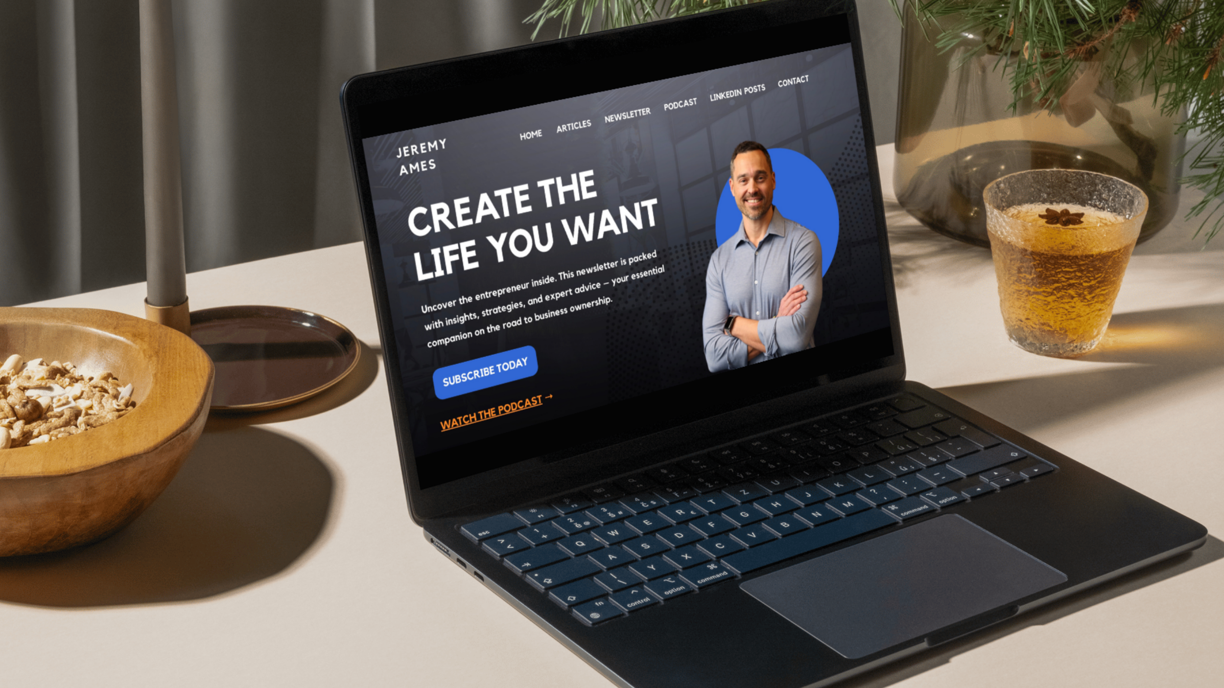 A laptop on a white table displaying a website titled 'Create the Life You Want' featuring a photo of a smiling man in a gray shirt. There are two bowls, one with grains and the other appears to have a drink, and a potted plant nearby.