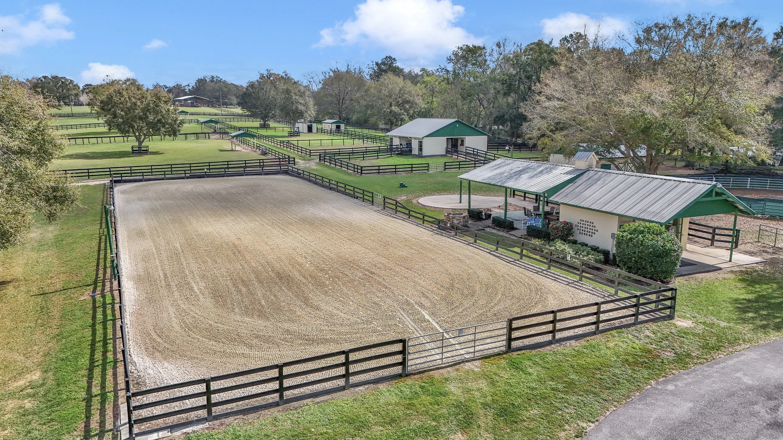 A large outdoor equestrian riding arena with fleet footing, surrounded by black wooden fences, and nearby shelters with green metal roofs. There's a grassy area with trees and additional fencing in the background, under a partly cloudy sky.