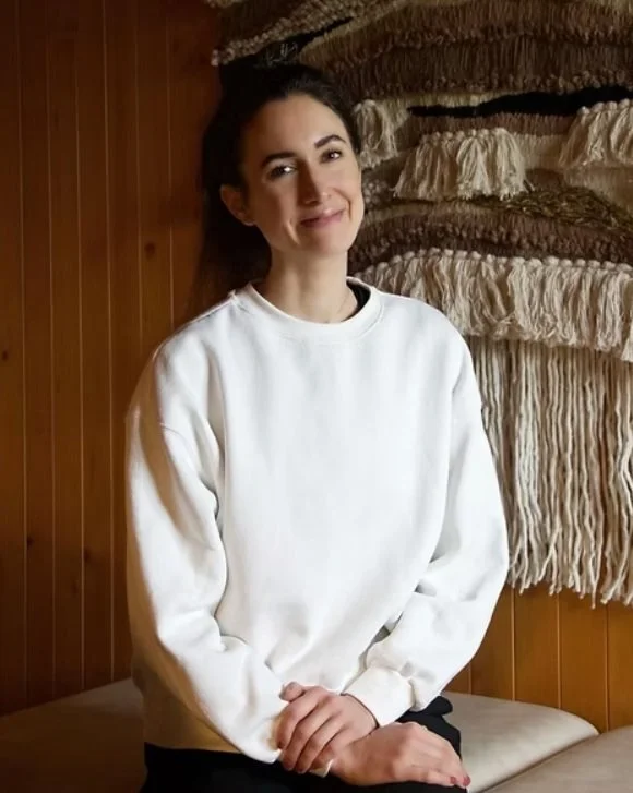 Une femme souriante porte un sweat blanc et est assise dans une pièce avec un mur en bois et un tapis mural en tissu coloré derrière elle.