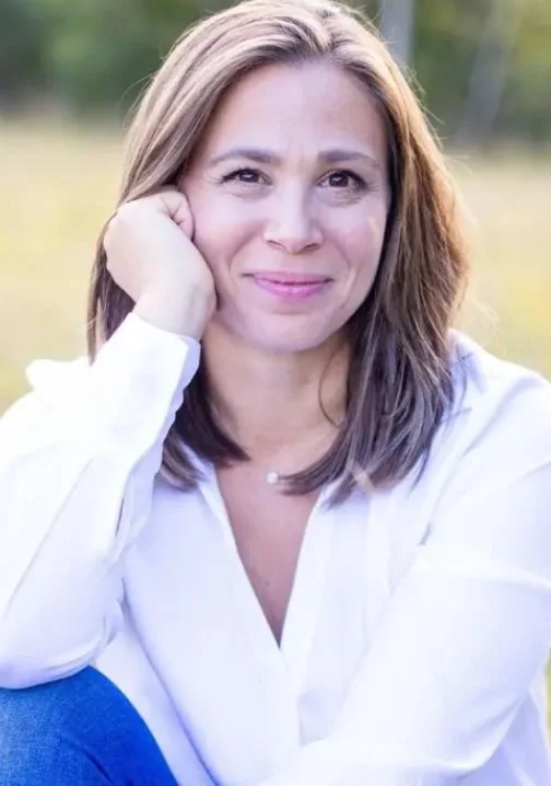 Une femme souriante avec les cheveux bruns, portant une chemise blanche, assise dans un champ en plein air.