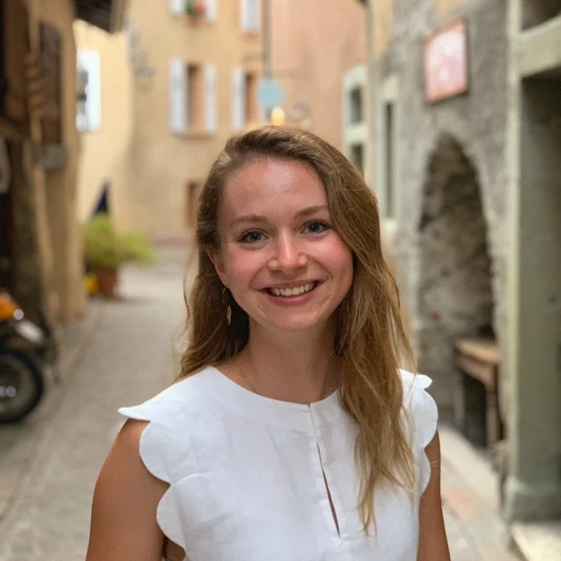 Jeune femme souriante avec des cheveux longs, portant une blouse blanche à manches volantes, dans une rue pavée ancienne avec des bâtiments en pierre.