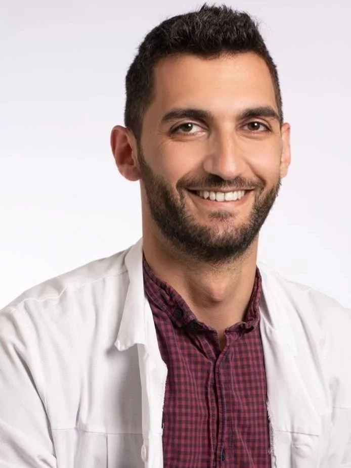 Un homme souriant, portant une blouse blanche et une chemise à carreaux rouge, fond blanc.