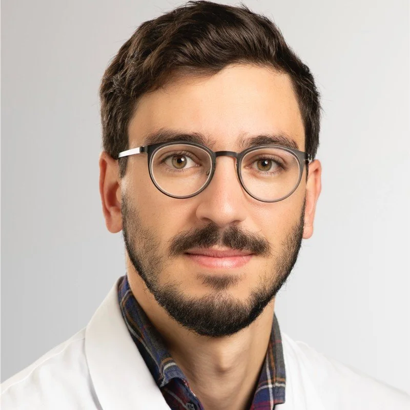 Un homme avec des lunettes, une barbe et un costume blanc, en gros plan, regardant l'objectif.