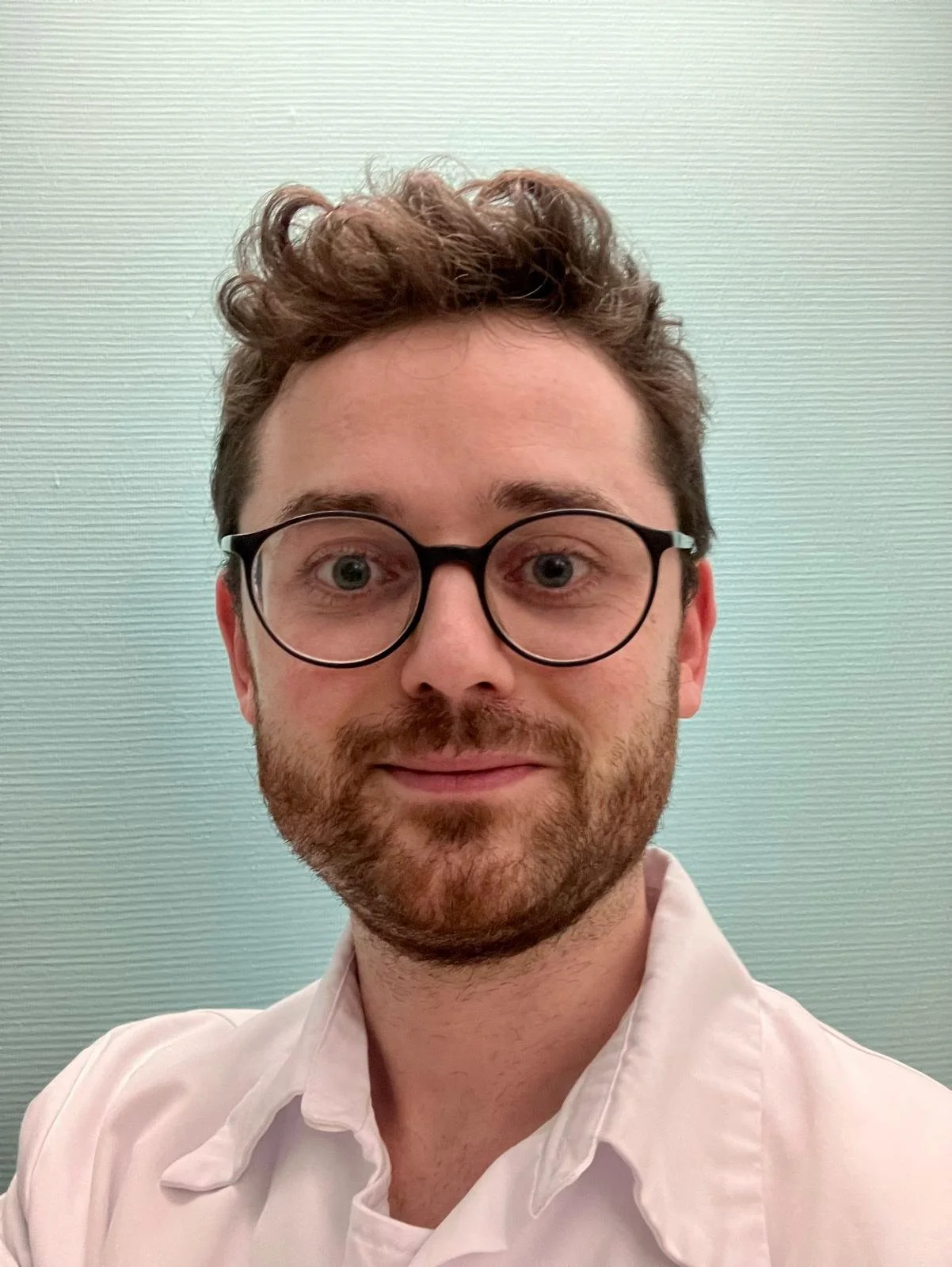 Un homme avec des cheveux bruns bouclés, portant des lunettes rondes, souriant, portant une chemise rose clair, contre un fond vert clair.