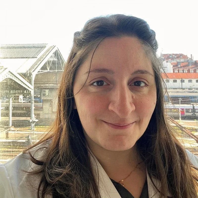 Jeune femme avec cheveux bruns longs, souriante, à proximité d'une fenêtre avec vue sur une gare ou un bâtiment historique, en journée.