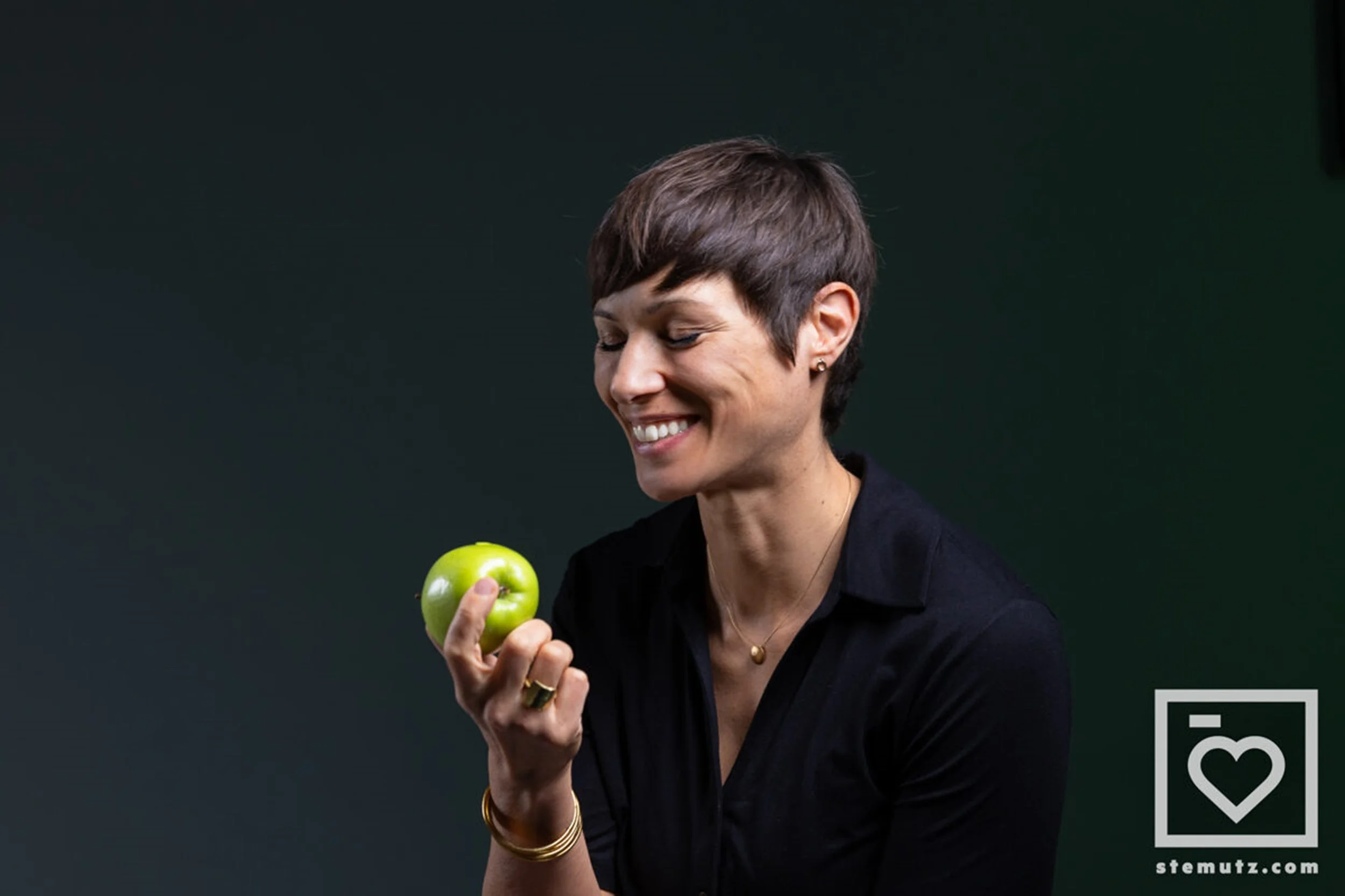 Une femme souriante tient une pomme verte dans sa main, portant des bijoux dorés et une chemise noire, contre un fond sombre.