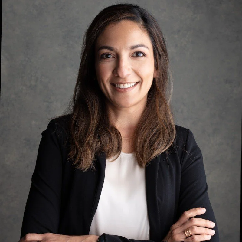 Une femme souriante avec des cheveux bruns, portant une veste noire et un chemisier blanc, se tient devant un fond gris.