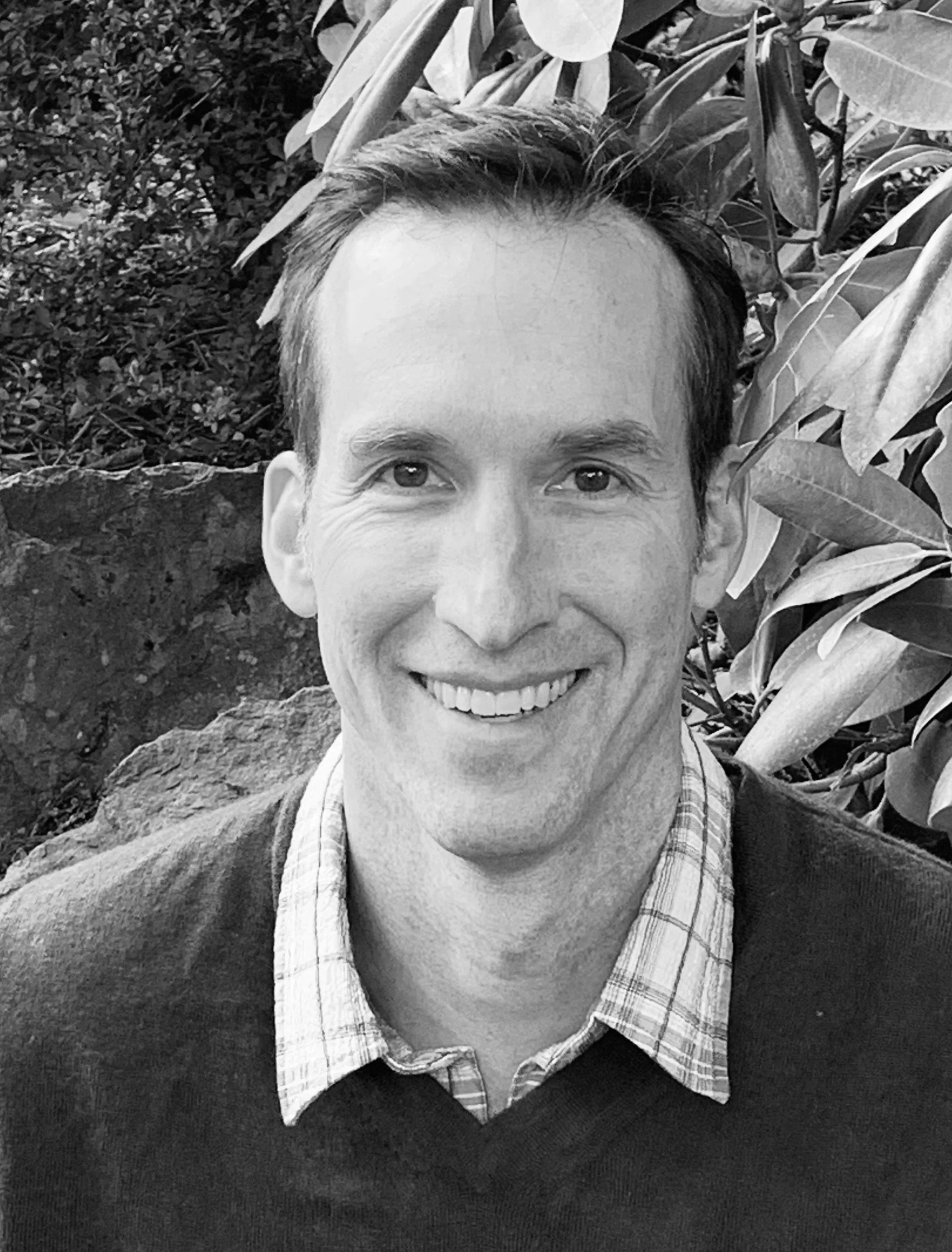 A black and white photo of a smiling man with short dark hair, wearing a checkered shirt and a sweater, outdoors with large leafy plants and a rock in the background.