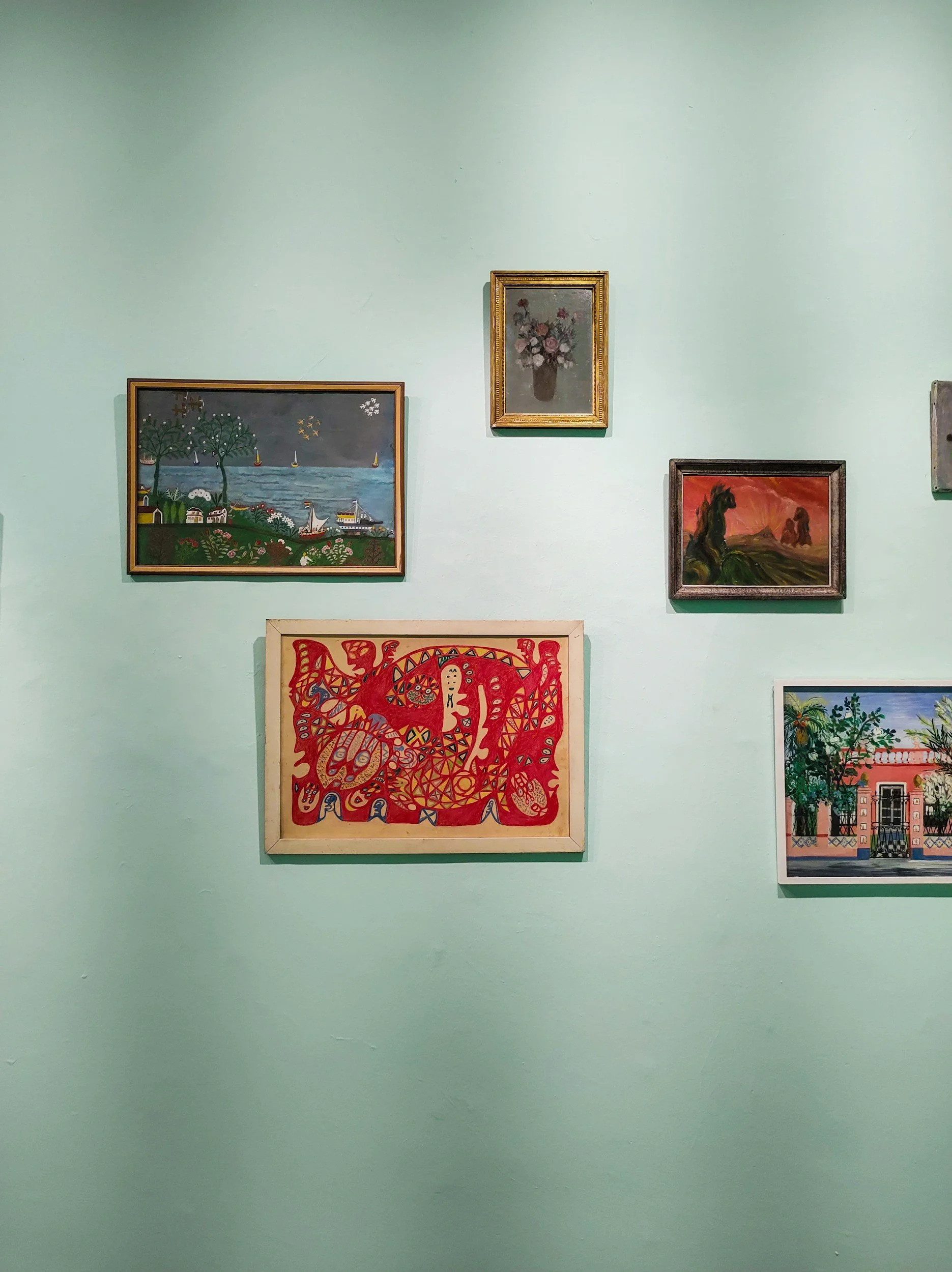 Collection of six framed paintings displayed on a light green wall, including landscape, floral, and abstract artworks.