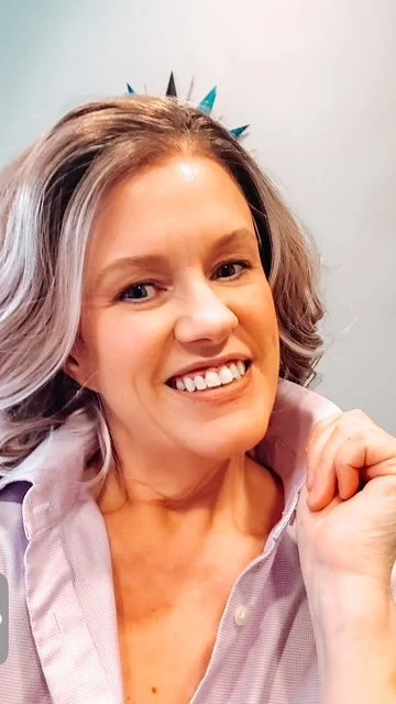 A woman with shoulder-length wavy blonde and light purple highlights wig, smiling, wearing a light purple collared shirt, and holding her fist near her chin. There is a plain light gray background.