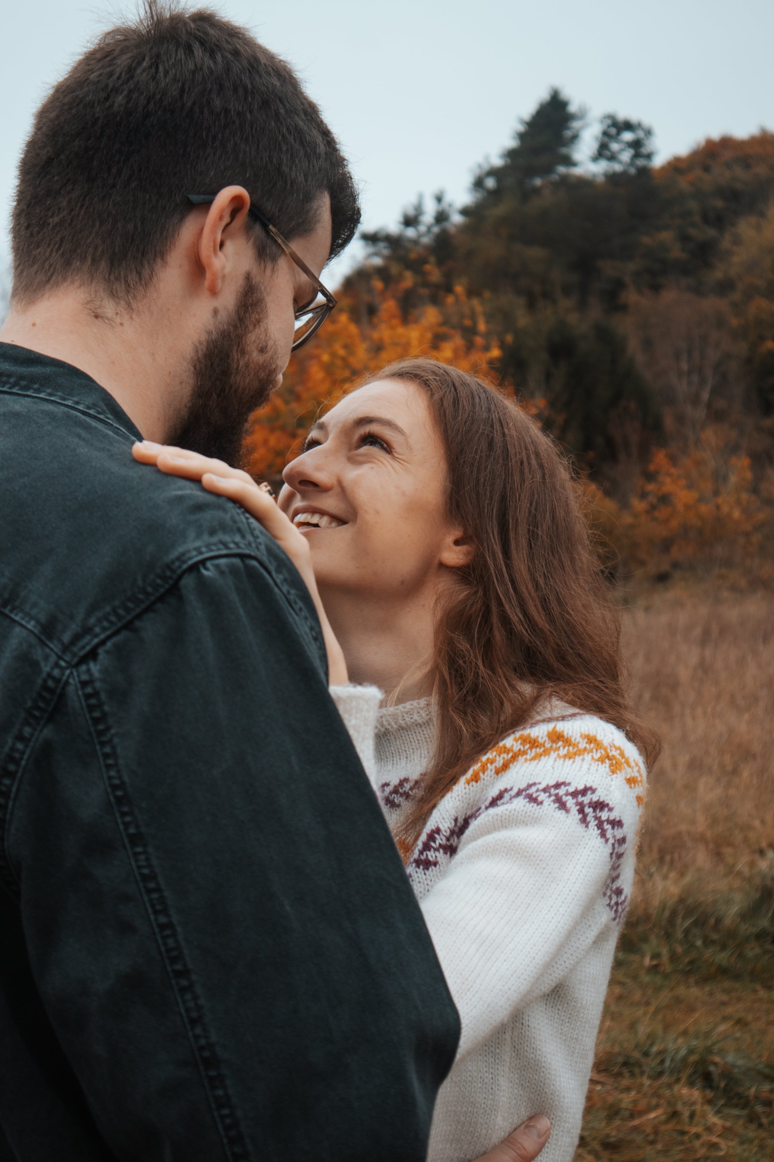 A couple standing close together outdoors in autumn, looking into each other's eyes with affection, with trees with fall foliage in the background.