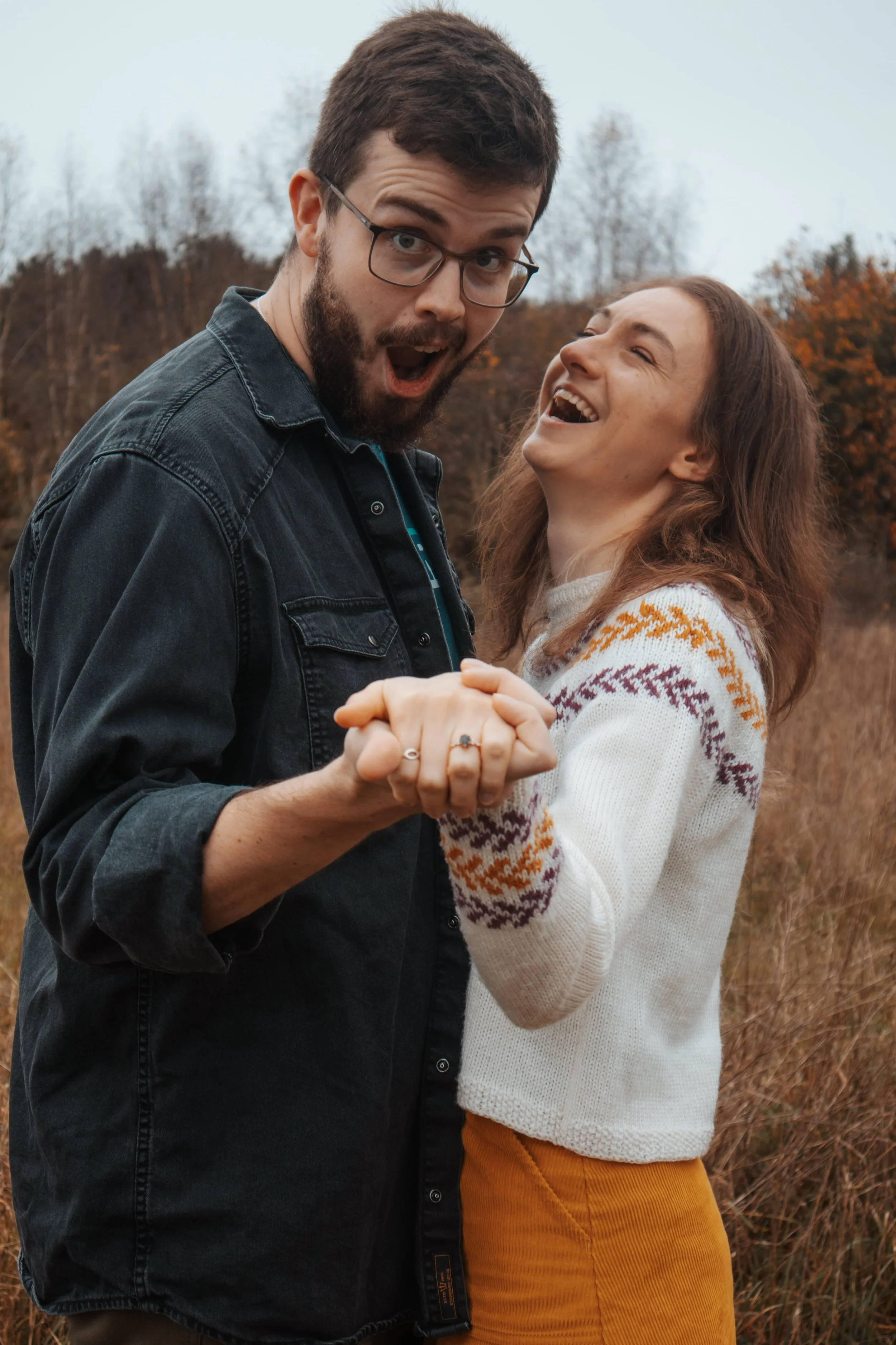 A couple holding hands, smiling and laughing outdoors in a field during autumn.